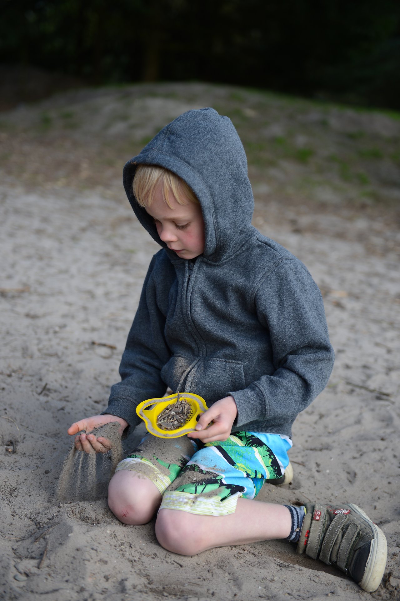 A child in a hoodie sits on the sand, letting it fall through their fingers while holding a yellow sieve.