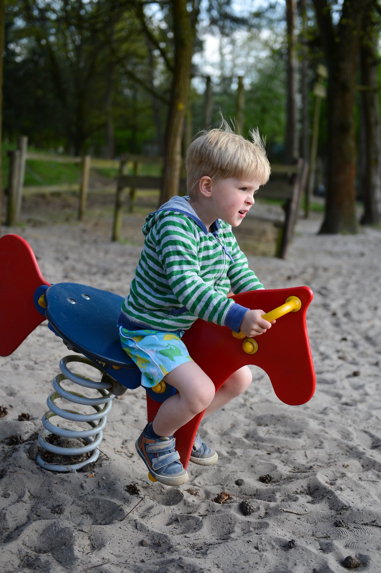 A young child in a striped hoodie rides a red spring rocker at a sandy playground.