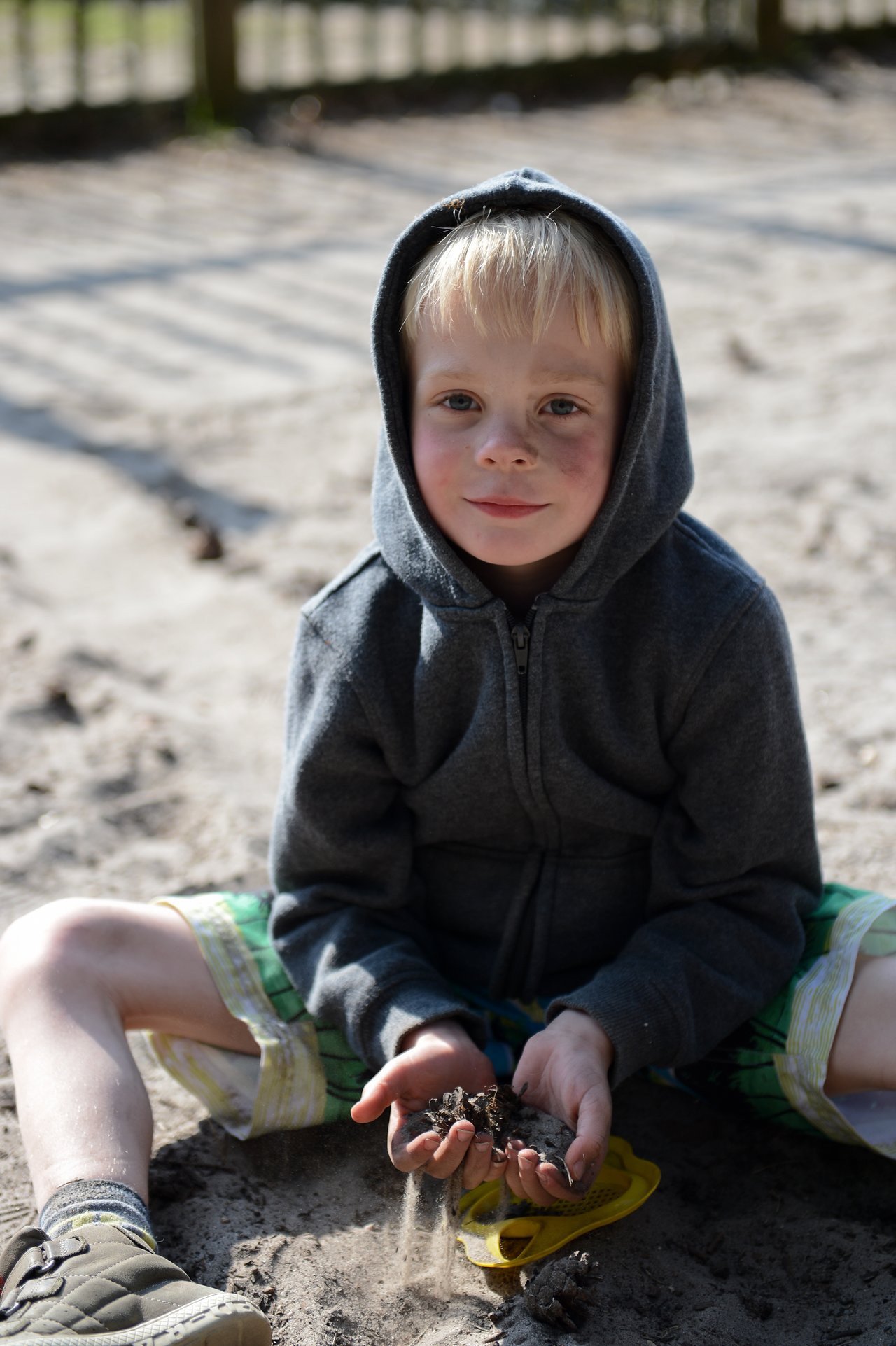 A child in a hoodie sits in the sand, holding dirt and pinecones while looking at the camera.