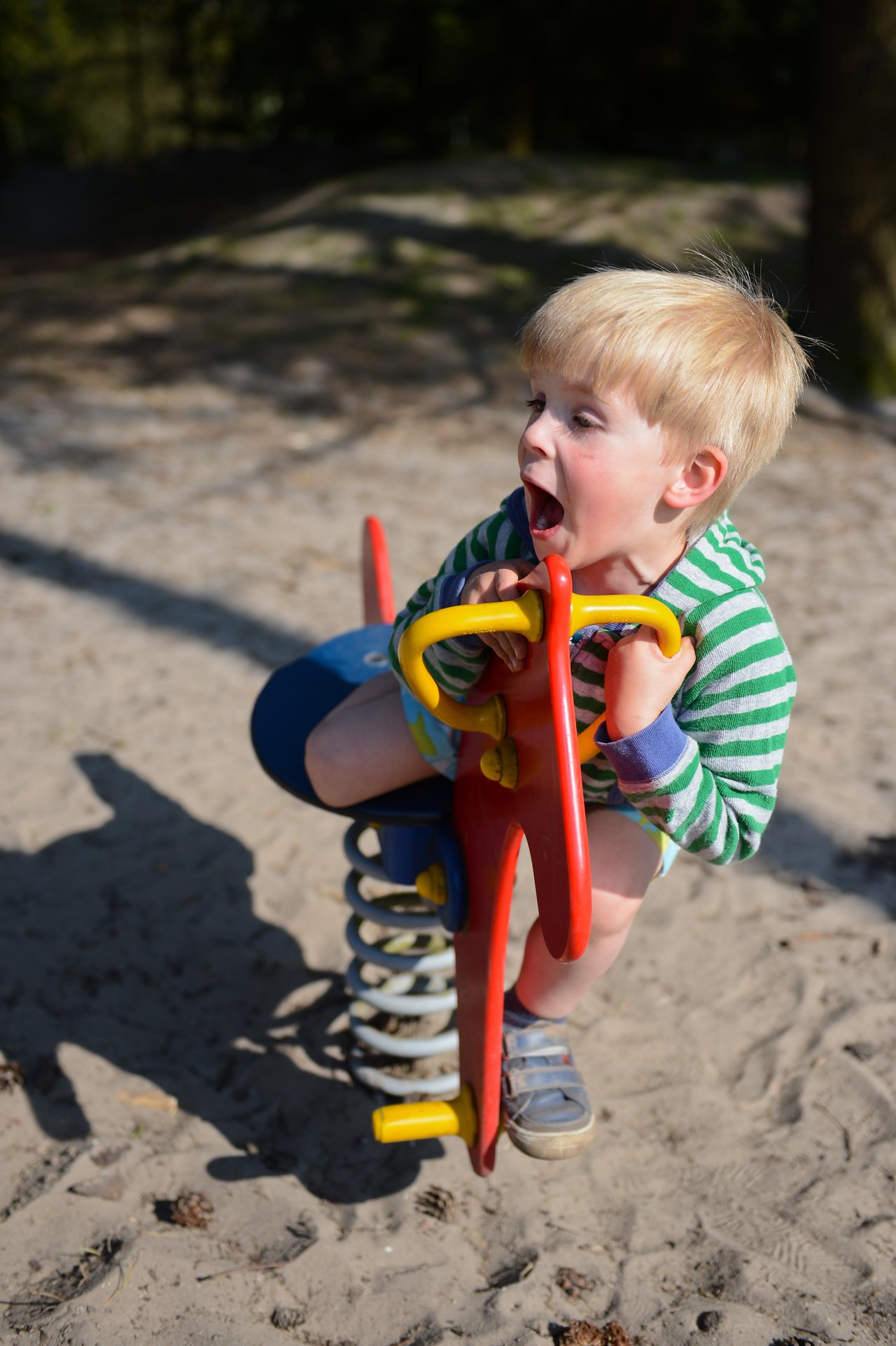 A young child in a striped shirt plays on a spring rocker at a sandy playground, mouth open excitedly.