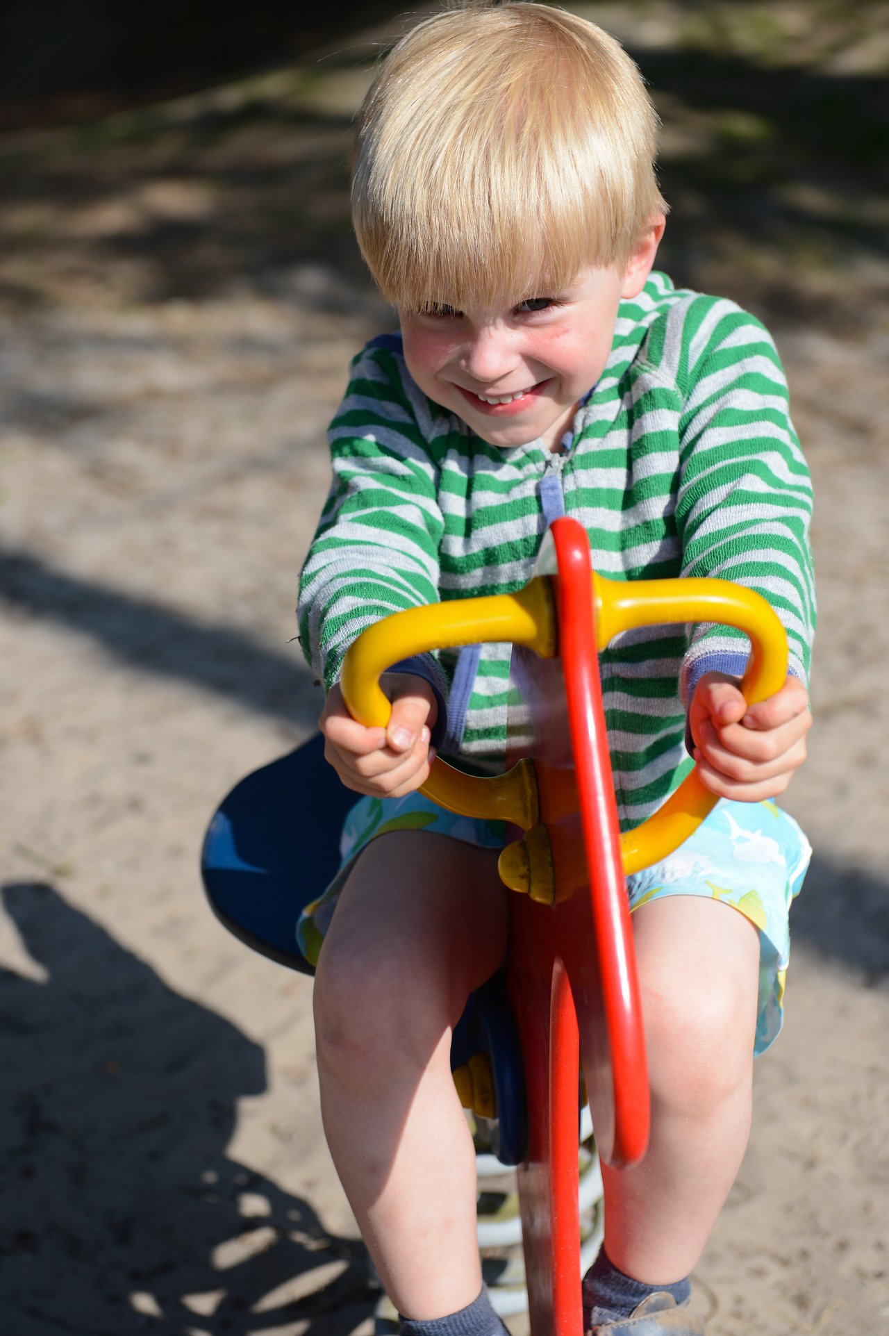 A young child in a striped sweater rides a colorful spring rocker at a playground, smiling.
