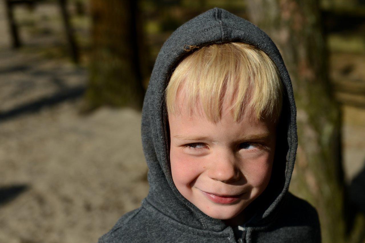 A young child in a gray hoodie smiles and looks to the side while standing outdoors in sunlight.