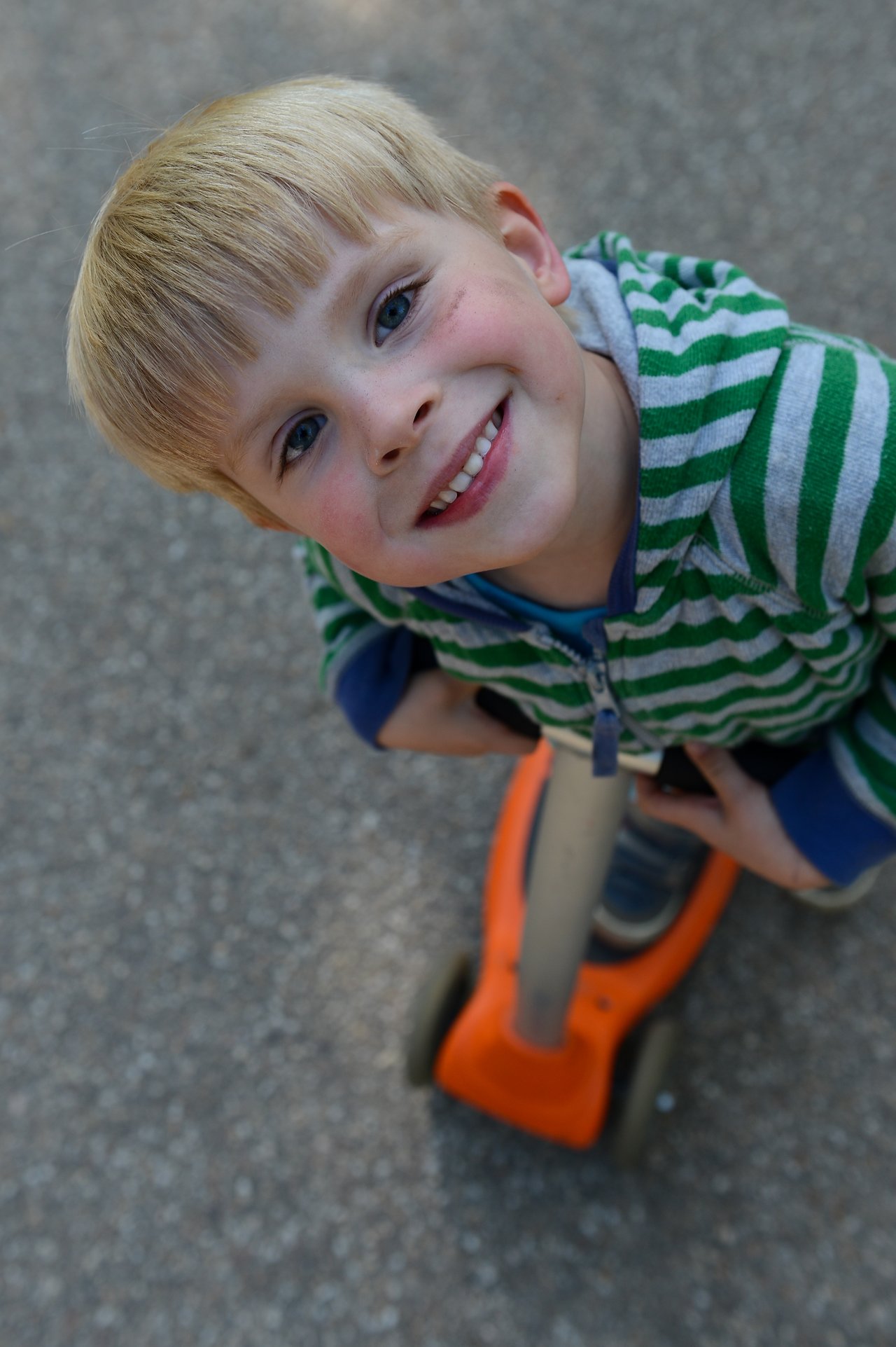 A smiling child in a green striped hoodie holds onto an orange scooter while looking up at the camera.