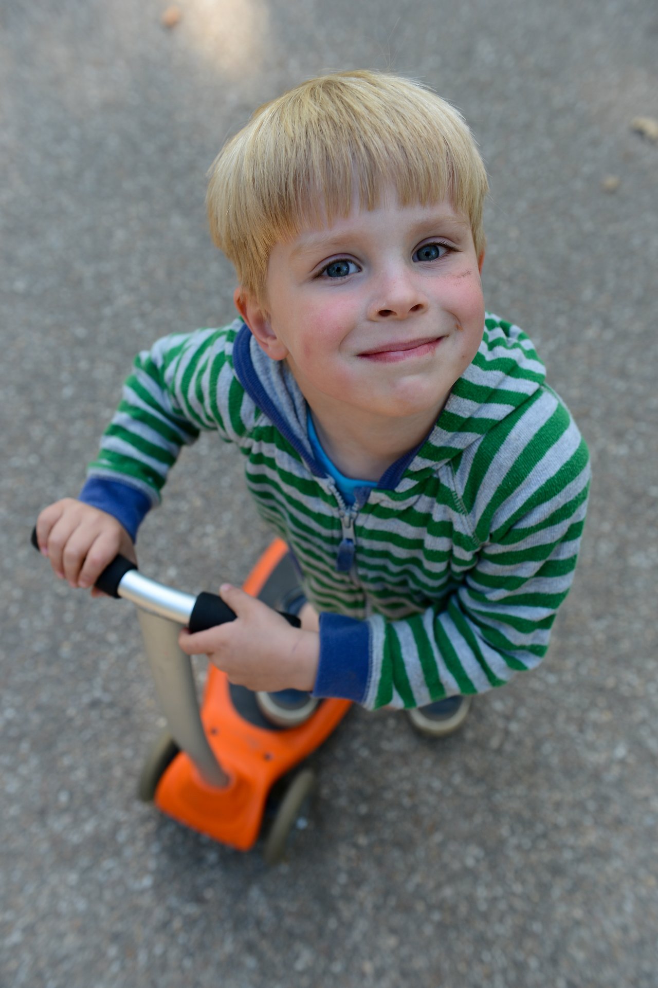 A young child in a striped hoodie smiles while holding the handlebars of an orange scooter on a paved path.