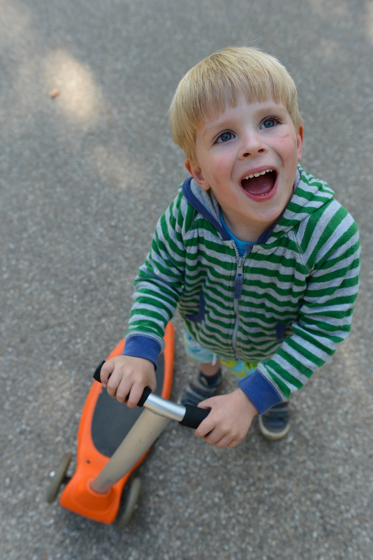 A smiling child in a striped hoodie holds the handlebars of an orange scooter while looking up.