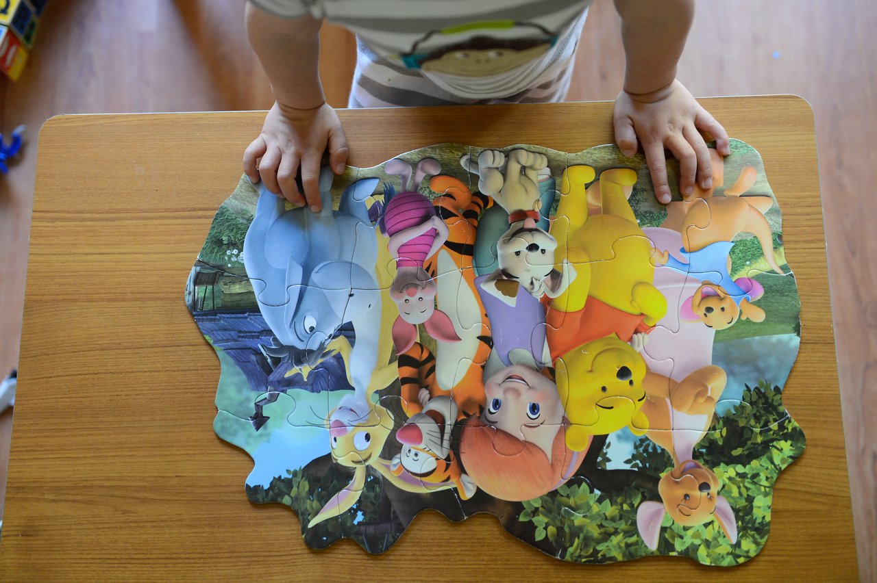 A child stands at a table, completing a colorful puzzle featuring Winnie the Pooh and friends.