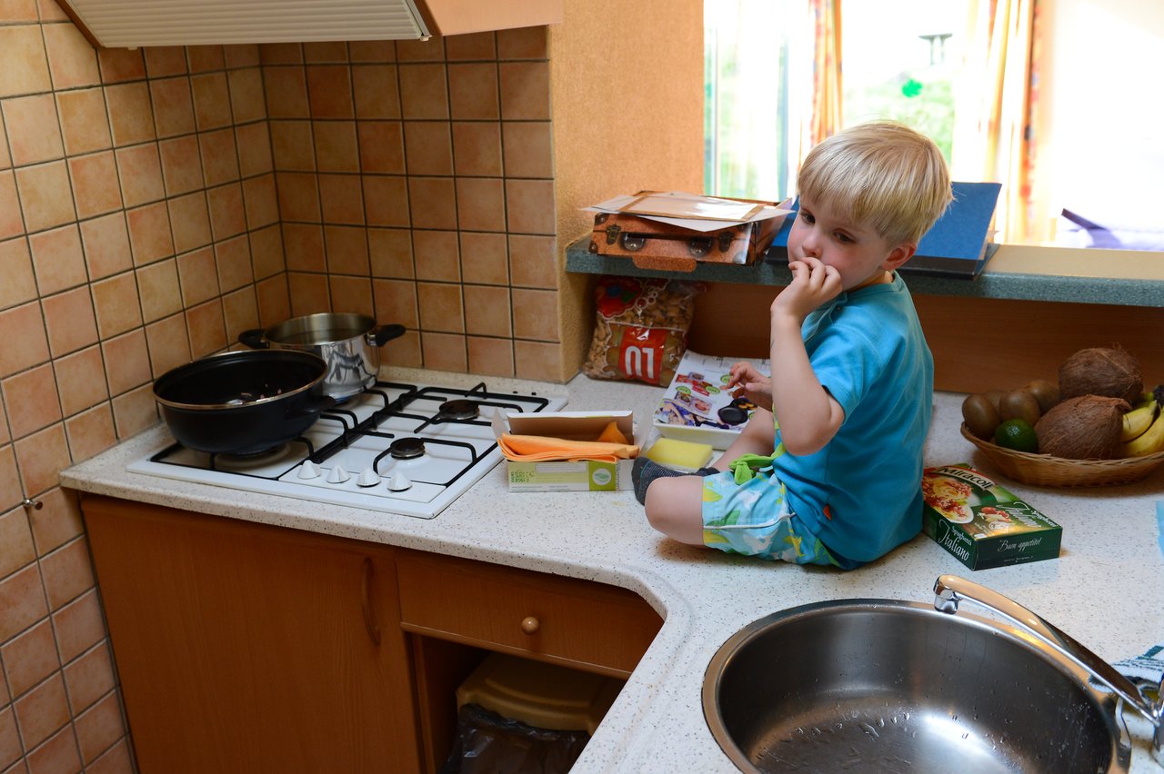 A young child sits on the kitchen counter, eating while surrounded by food items near a stove.