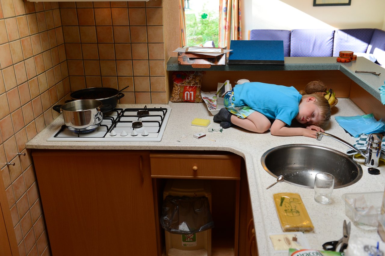 A young child in a blue shirt is lying curled up on a kitchen counter near the sink.