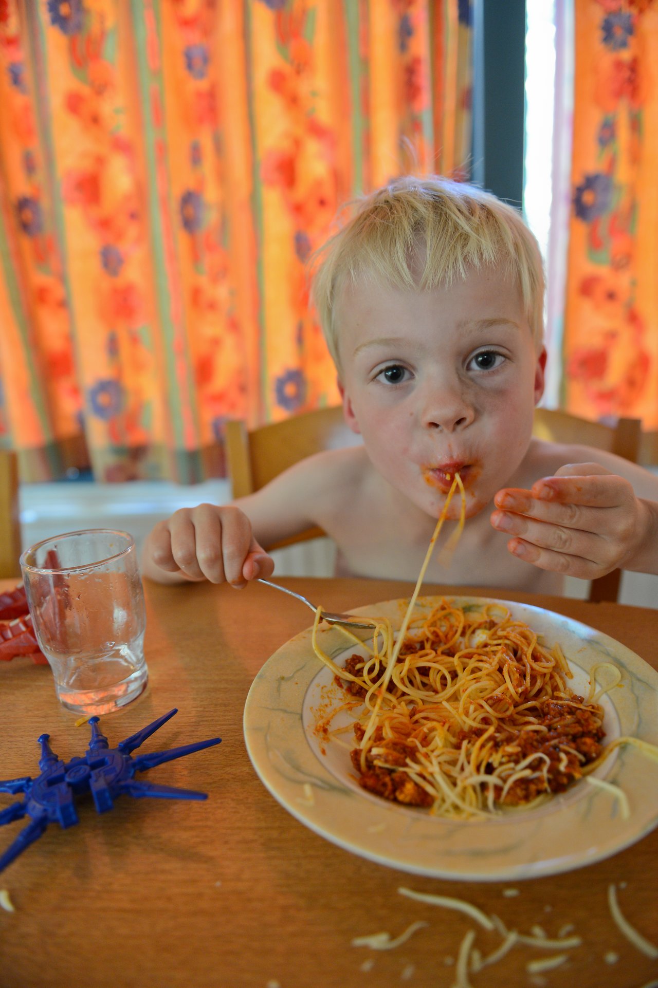 A young child eats spaghetti with their hands and a fork, sitting at a table with a toy nearby.
