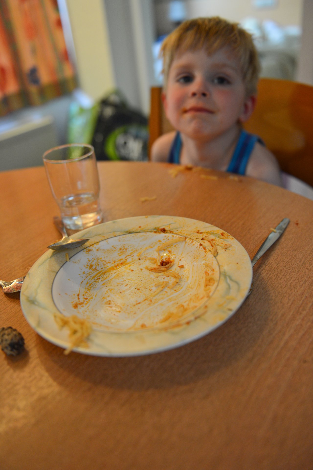 A young child with a messy face sits at a table with an empty plate of spaghetti.