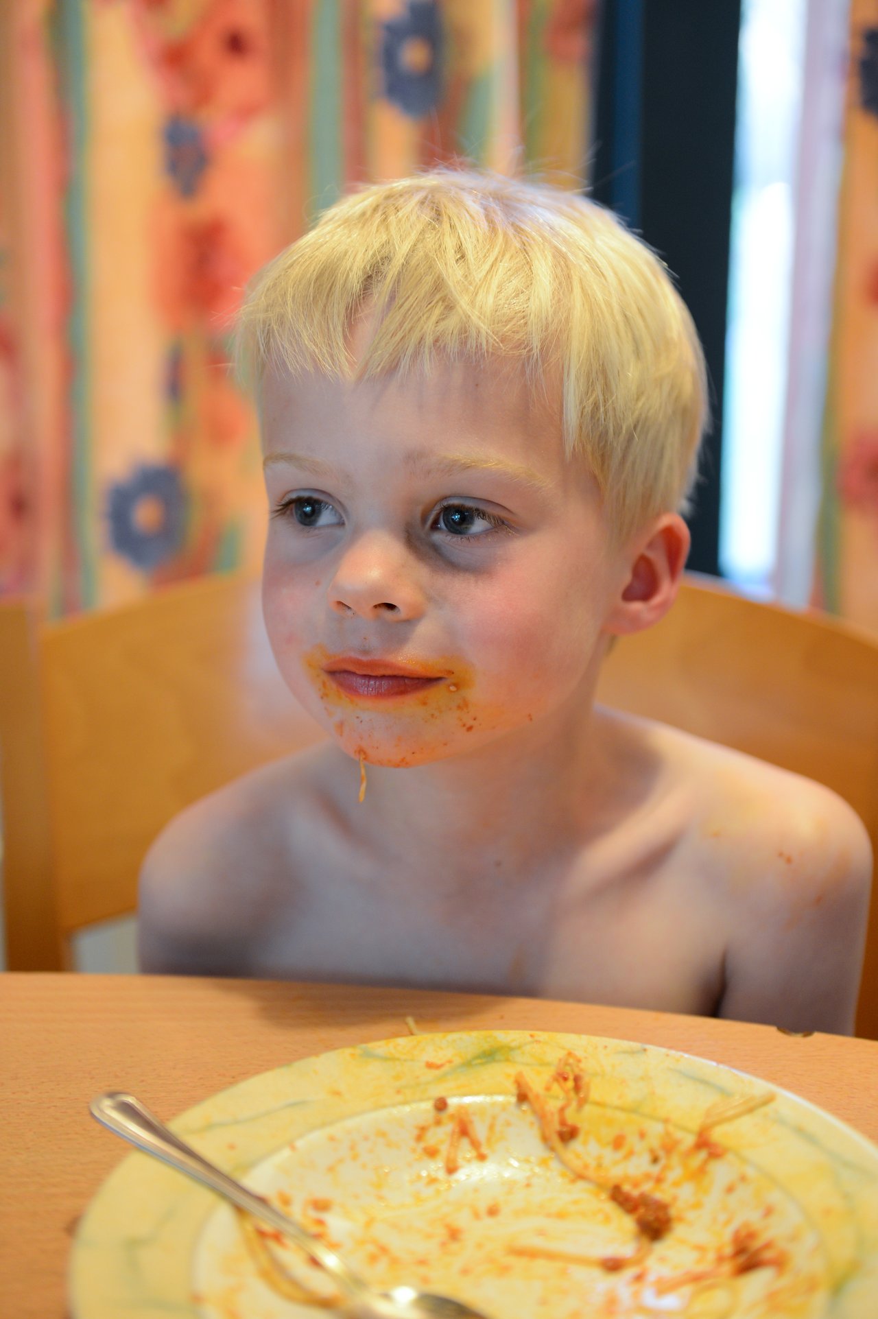 A young child with a messy face sits at a table after eating spaghetti, with an empty plate nearby.