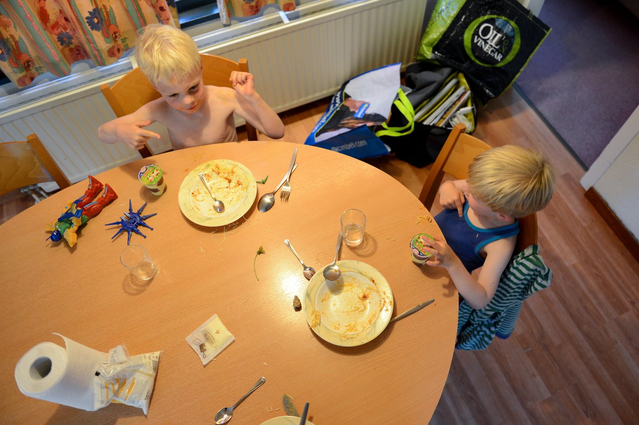 Two young children sit at a table with empty plates, one gesturing excitedly while the other eats from a cup.