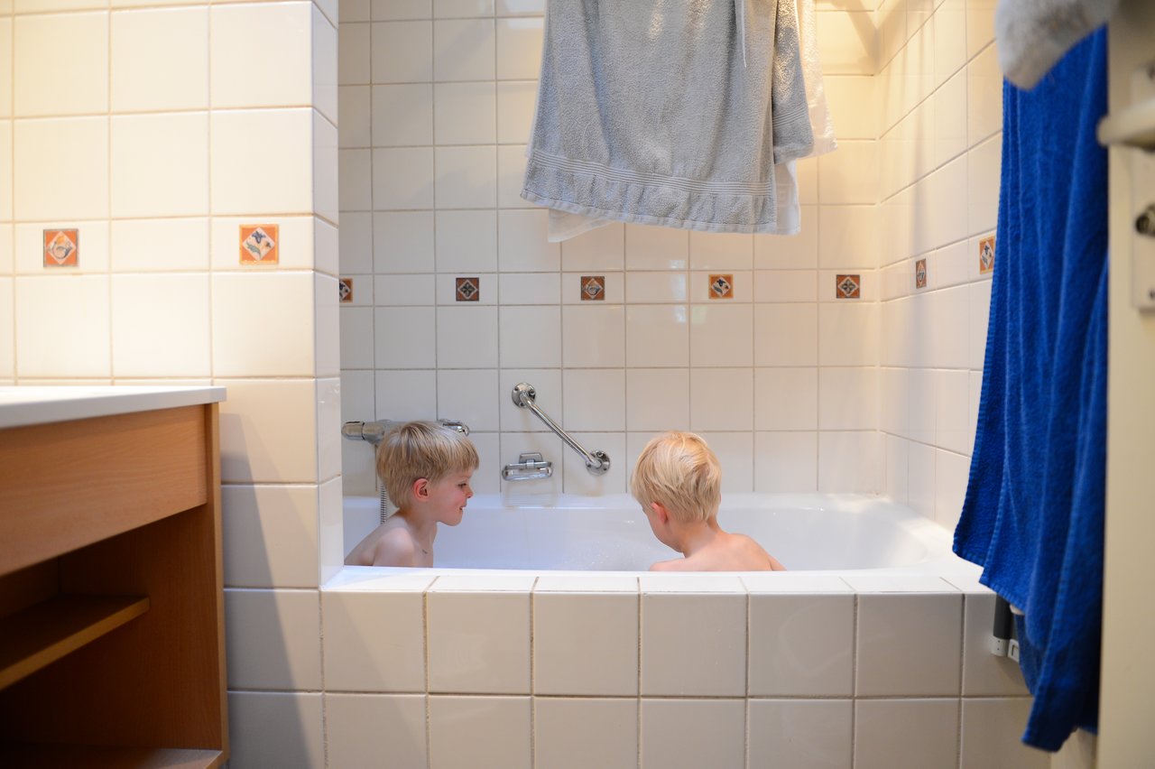 Two young children sit in a bathtub, facing each other and playing in the water.
