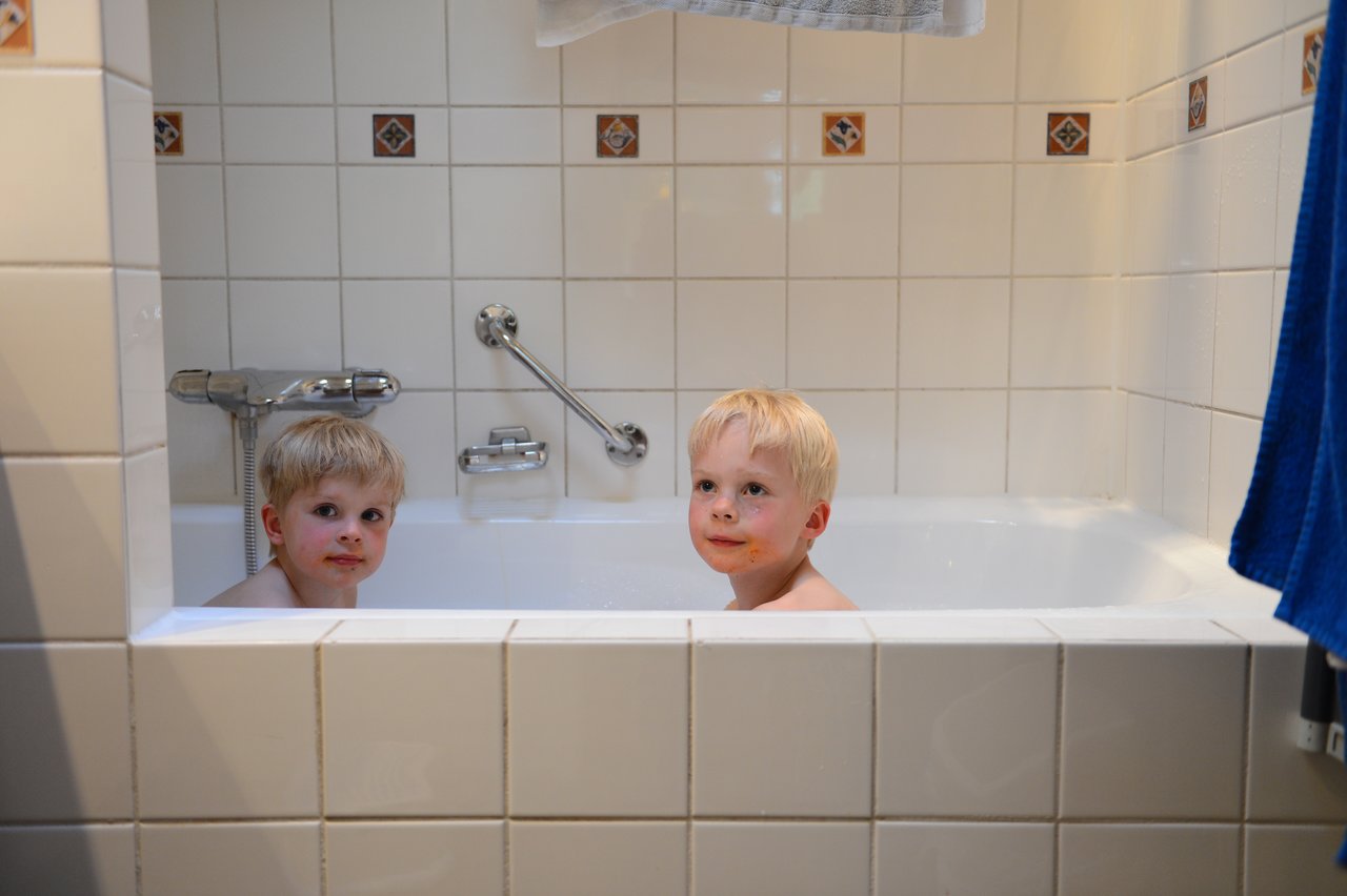 Two young children sit in a white bathtub, looking up with curious expressions.