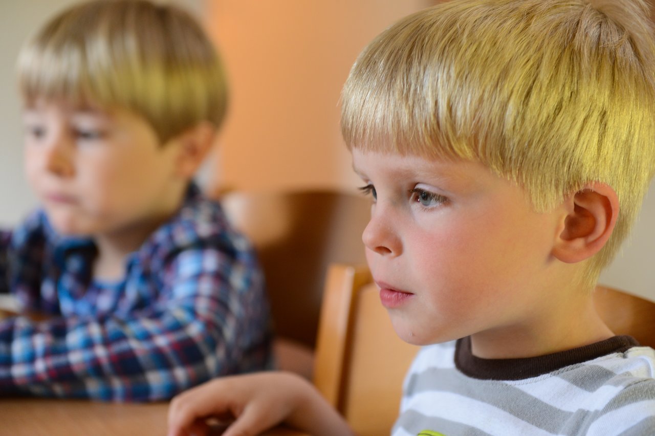 Two young boys with blond hair sit at a table, one in focus looking intently at something off-camera.