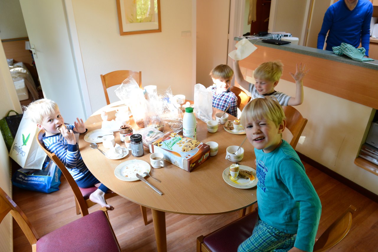 Four young children sit around a table with breakfast items, smiling and waving, while an adult stands nearby.