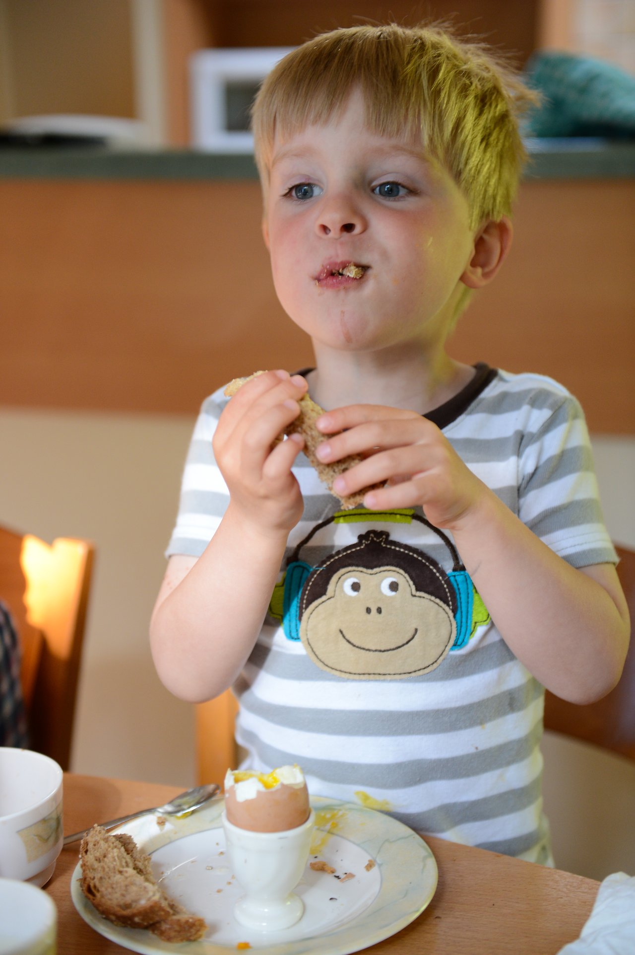 A young child eats bread at a table with a soft-boiled egg and dishes in front of them.