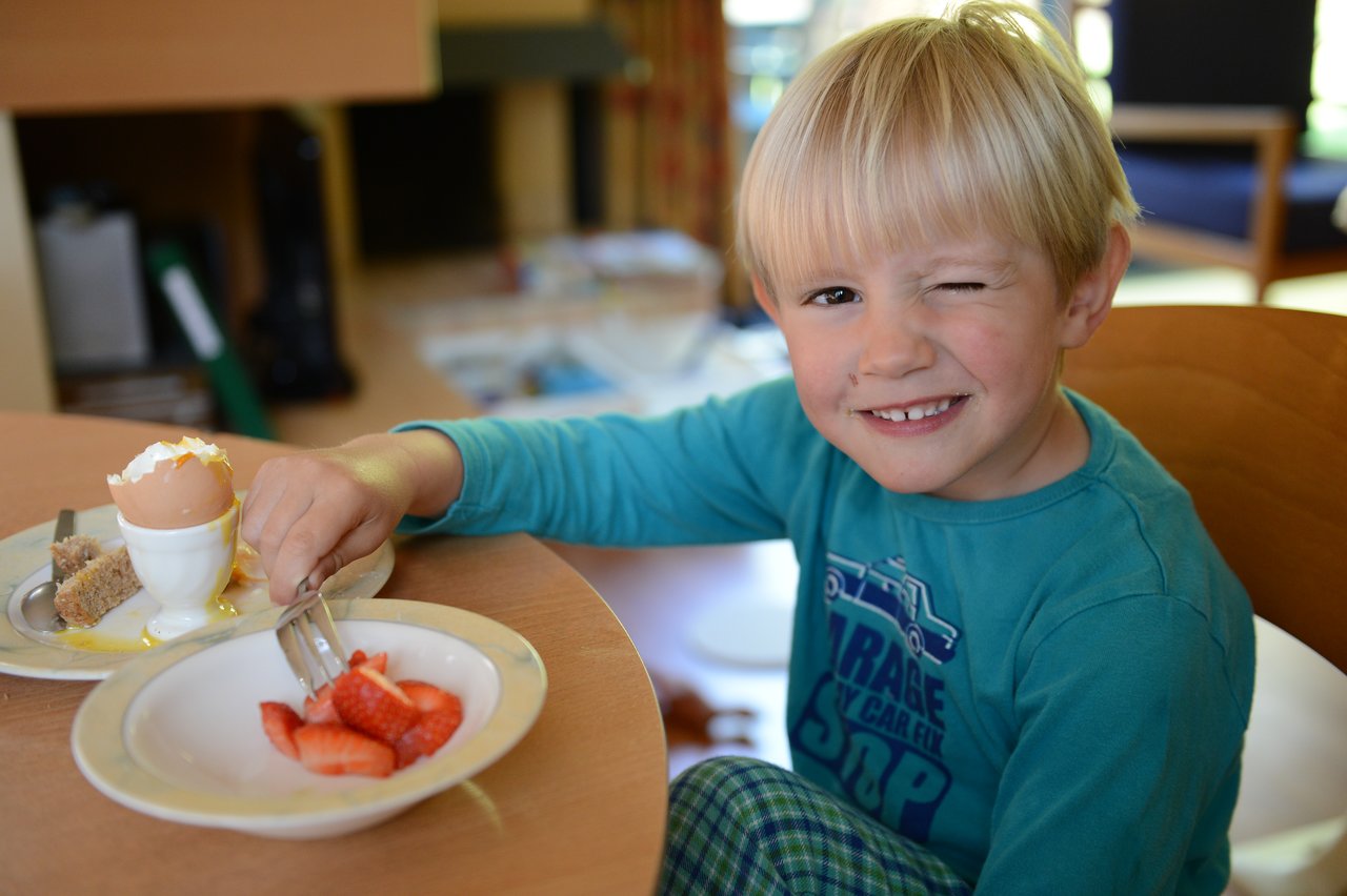 A young child in a blue shirt winks while eating breakfast with strawberries and a soft-boiled egg.