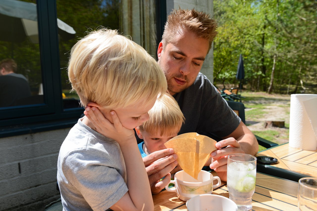 A man and two children pour liquid through a filter into a cup at an outdoor table.