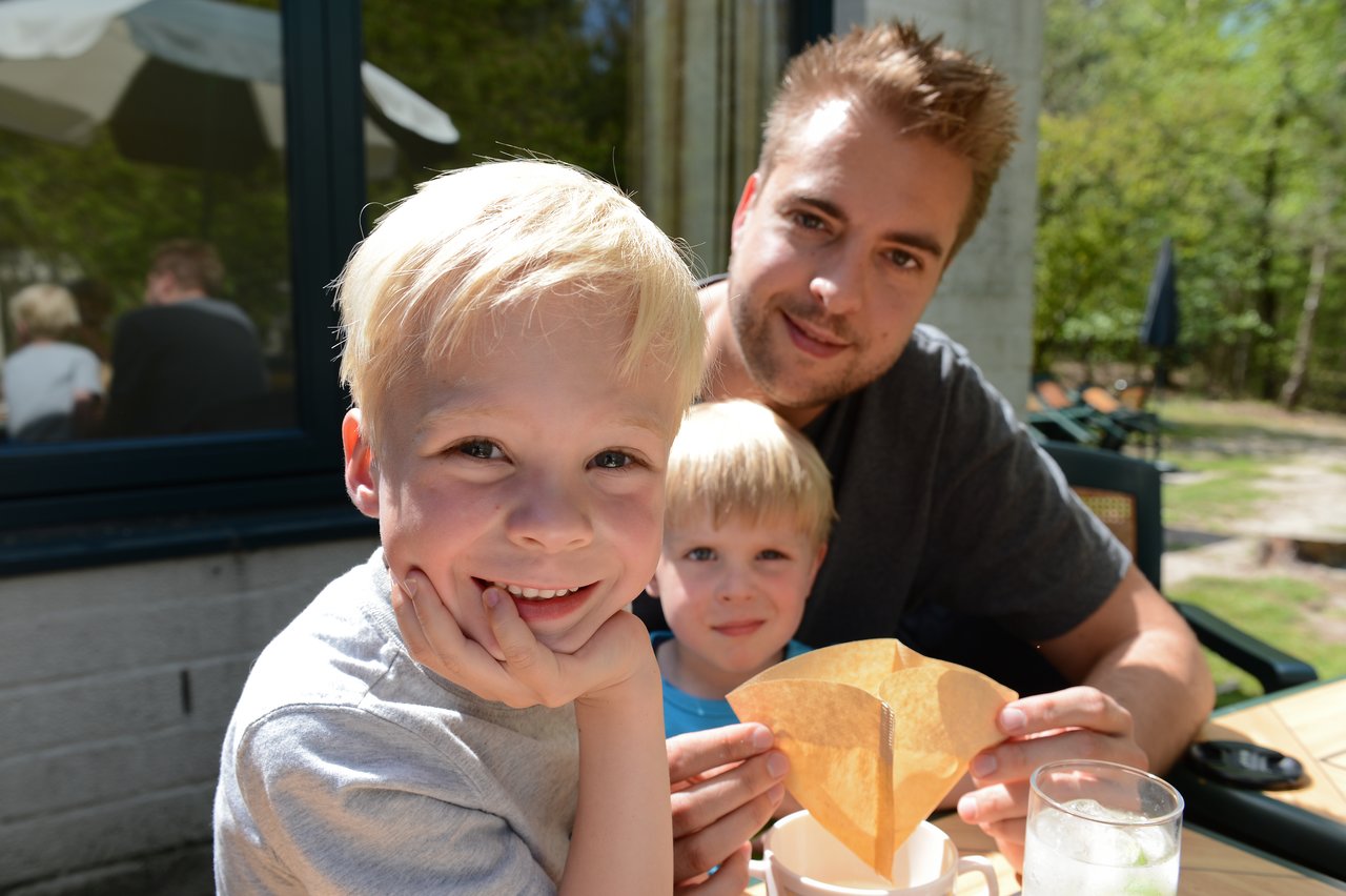 A smiling child in the foreground, with a man and another child sitting at an outdoor table behind him.