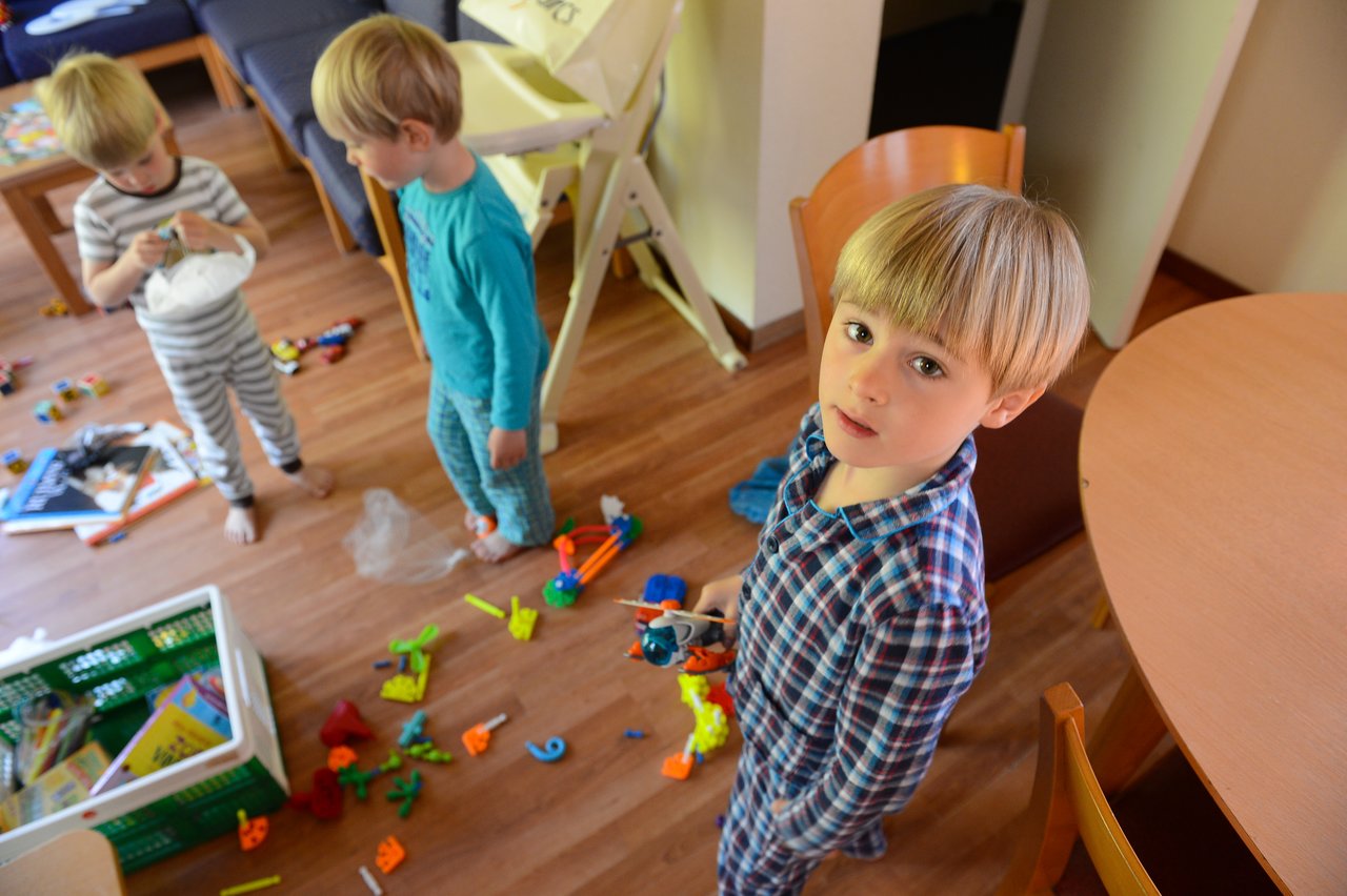 Three young children in pajamas play with toys on the floor, with one looking directly at the camera.