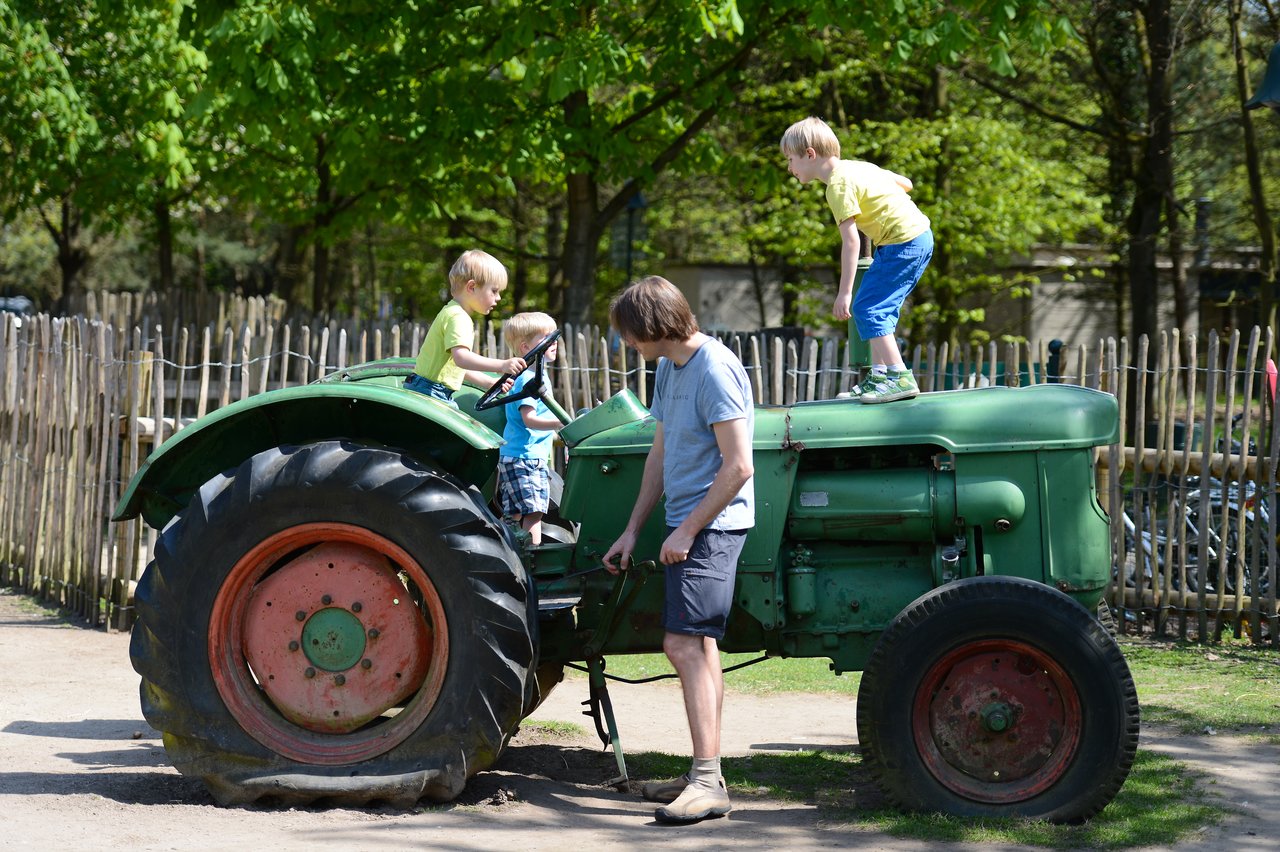 An adult helps children climb and play on an old green tractor in an outdoor setting.