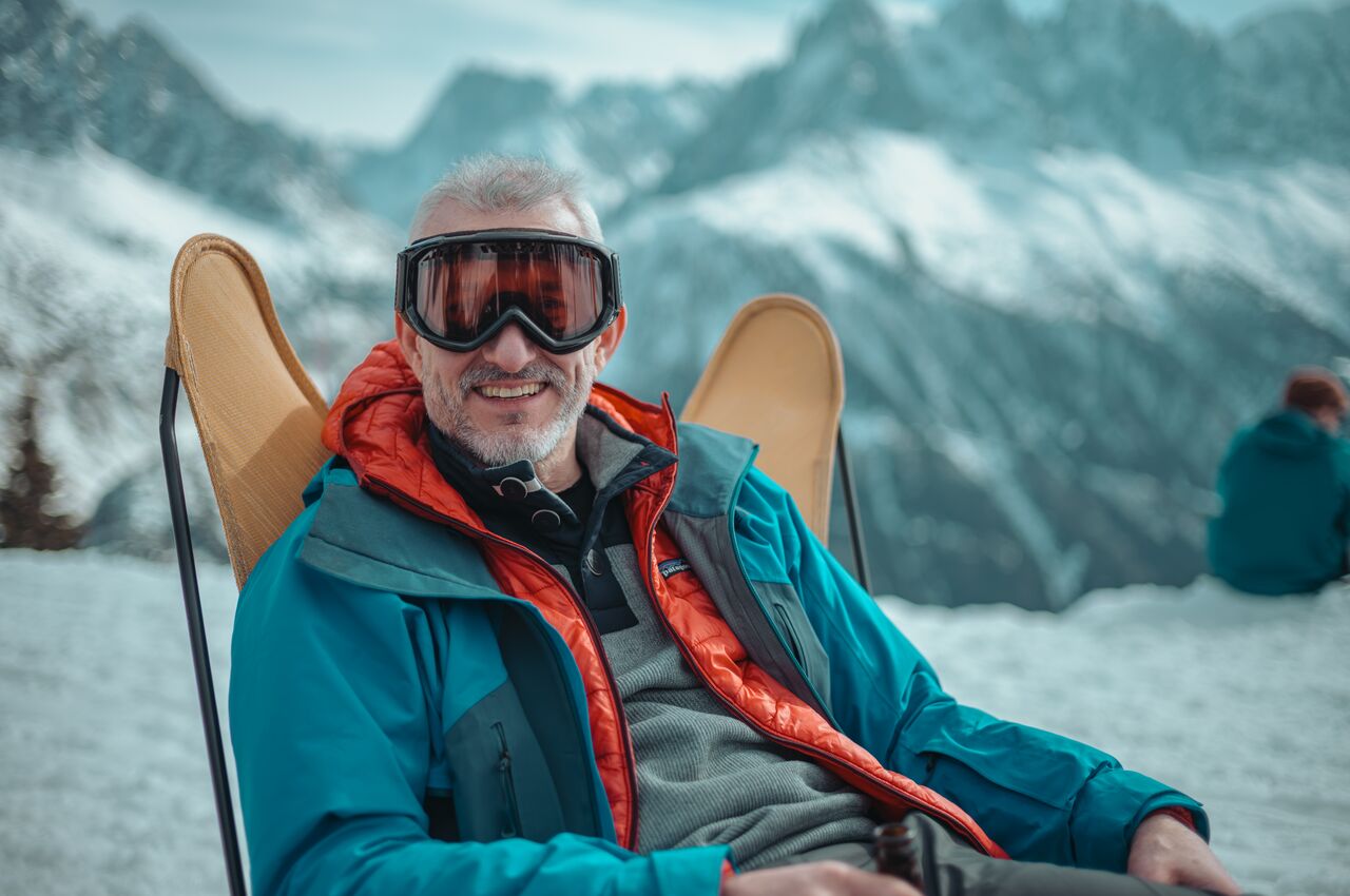 Smiling man in ski goggles relaxes in a lounge chair after skiing, holding a drink and bundled in winter layers.