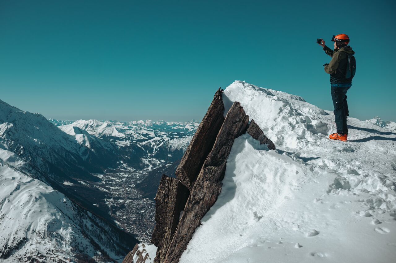 Person in orange helmet and ski boots stands on a snowy ridge at Bochard summit, photographing the Chamonix valley and surrounding snow-covered mountains.