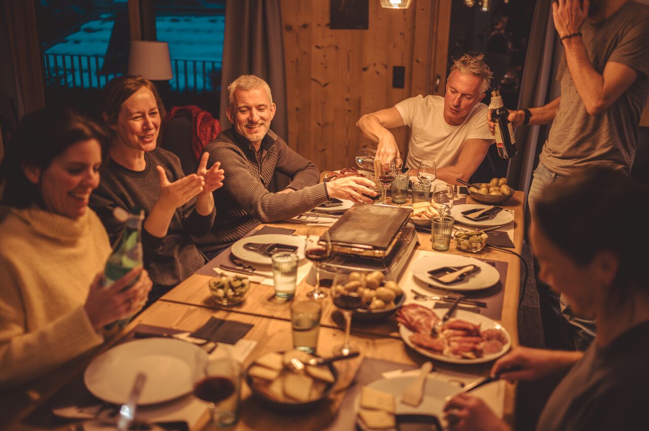 Six friends sit around a table, melting raclette cheese and pouring wine, laughing while serving potatoes and meats from shared plates.