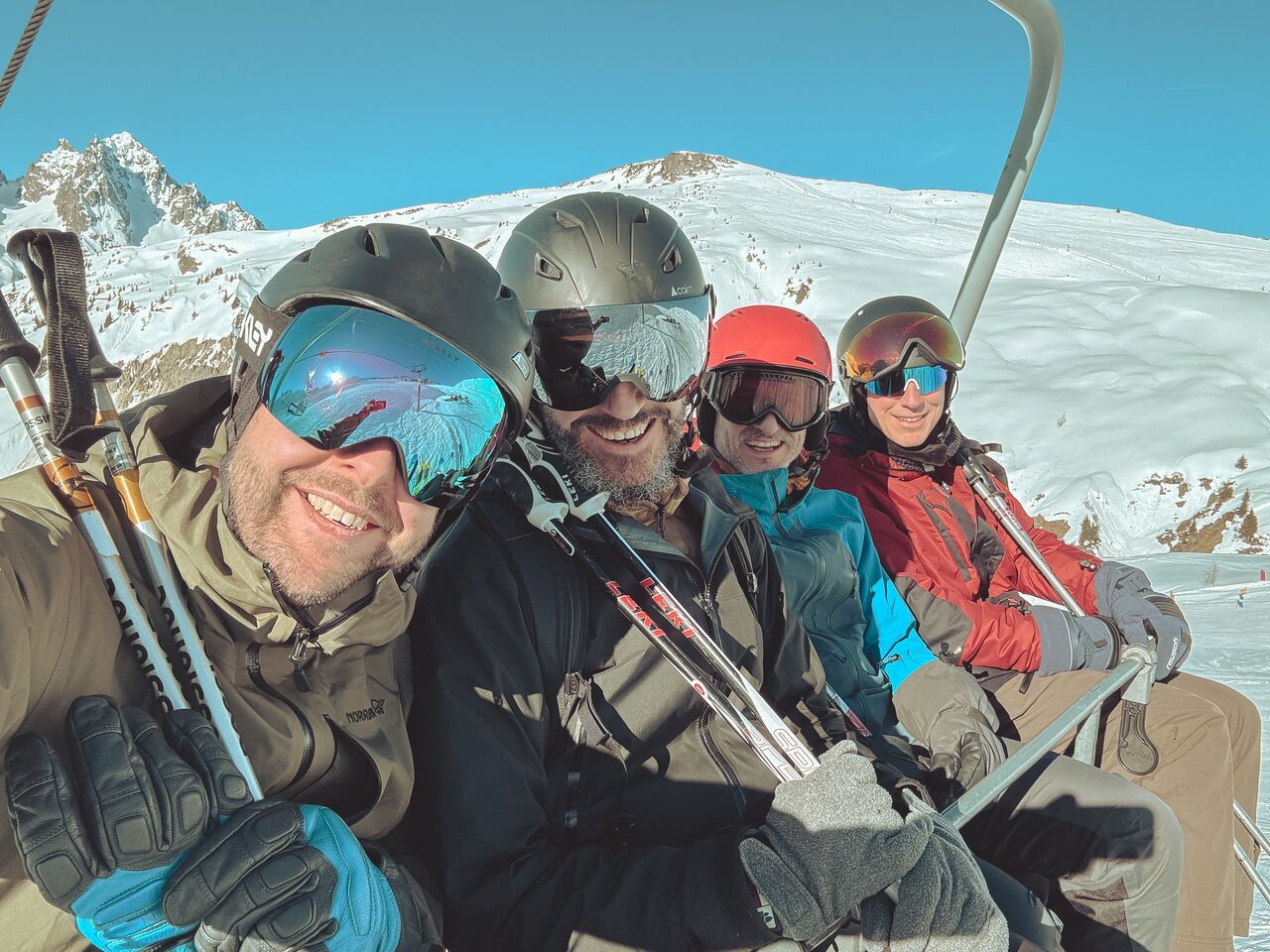 Four friends take a smiling selfie on a ski lift, wearing helmets and goggles, holding ski poles while riding above snowy mountain slopes.
