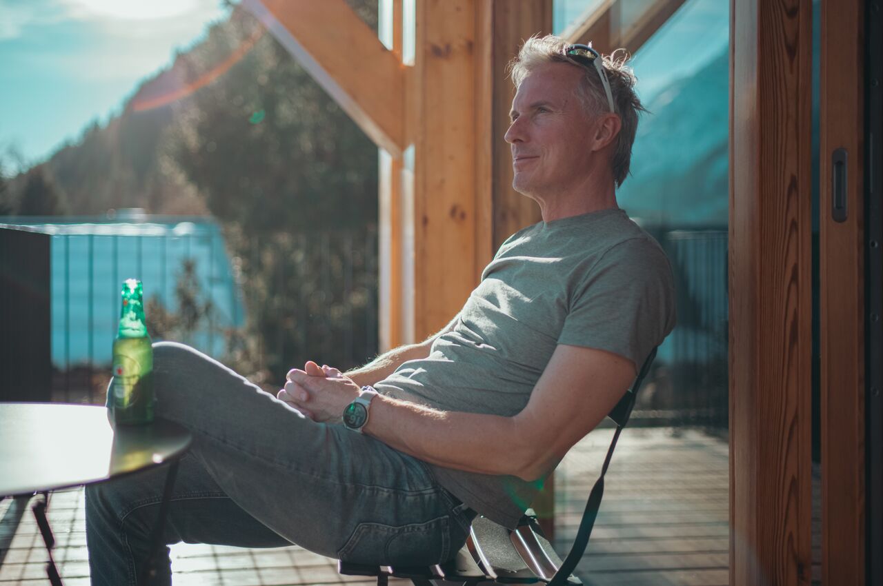 Man leans back on a sunny deck chair, hands clasped, sunglasses on his head, beside a small table with a beer bottle.