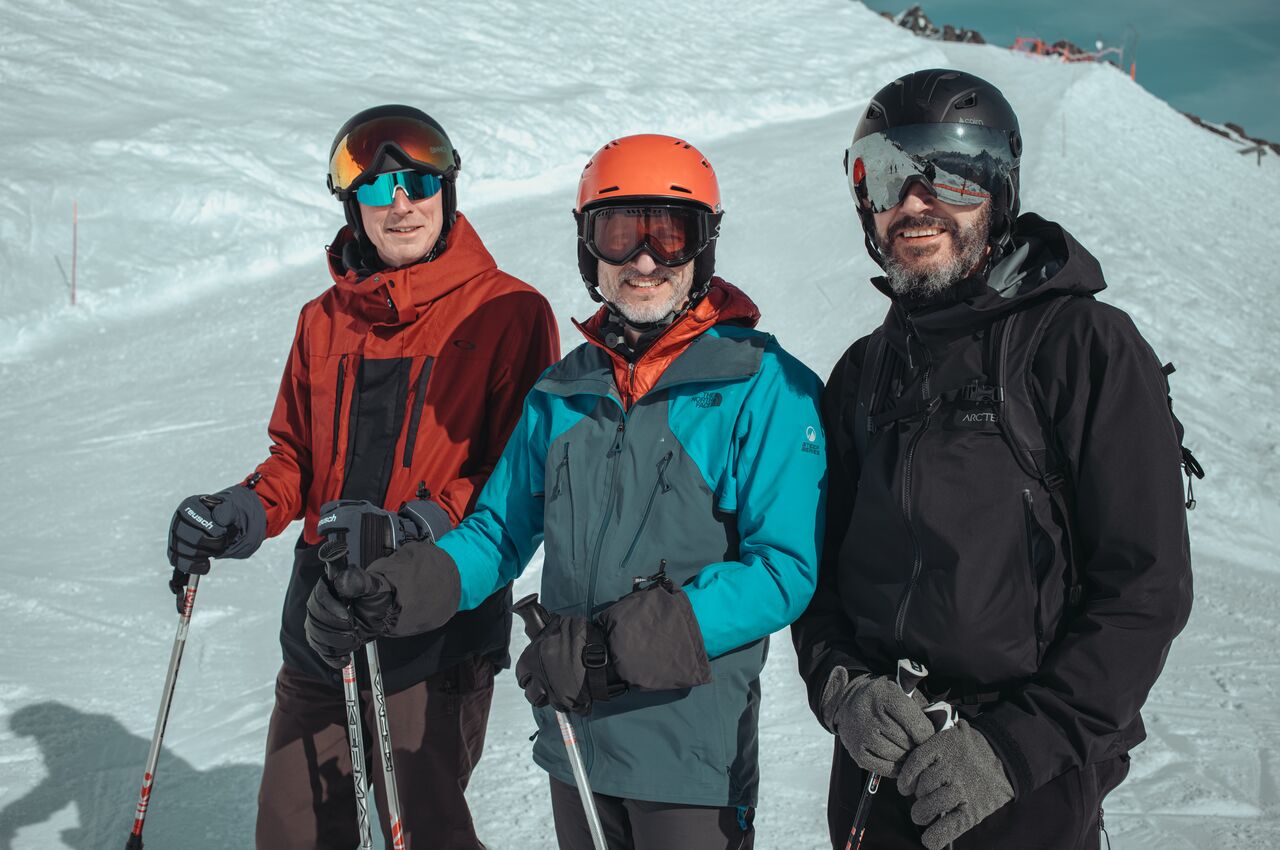 Three skiers wearing helmets and goggles stand together on a snowy slope, smiling at the camera and holding ski poles, about to continue their run.