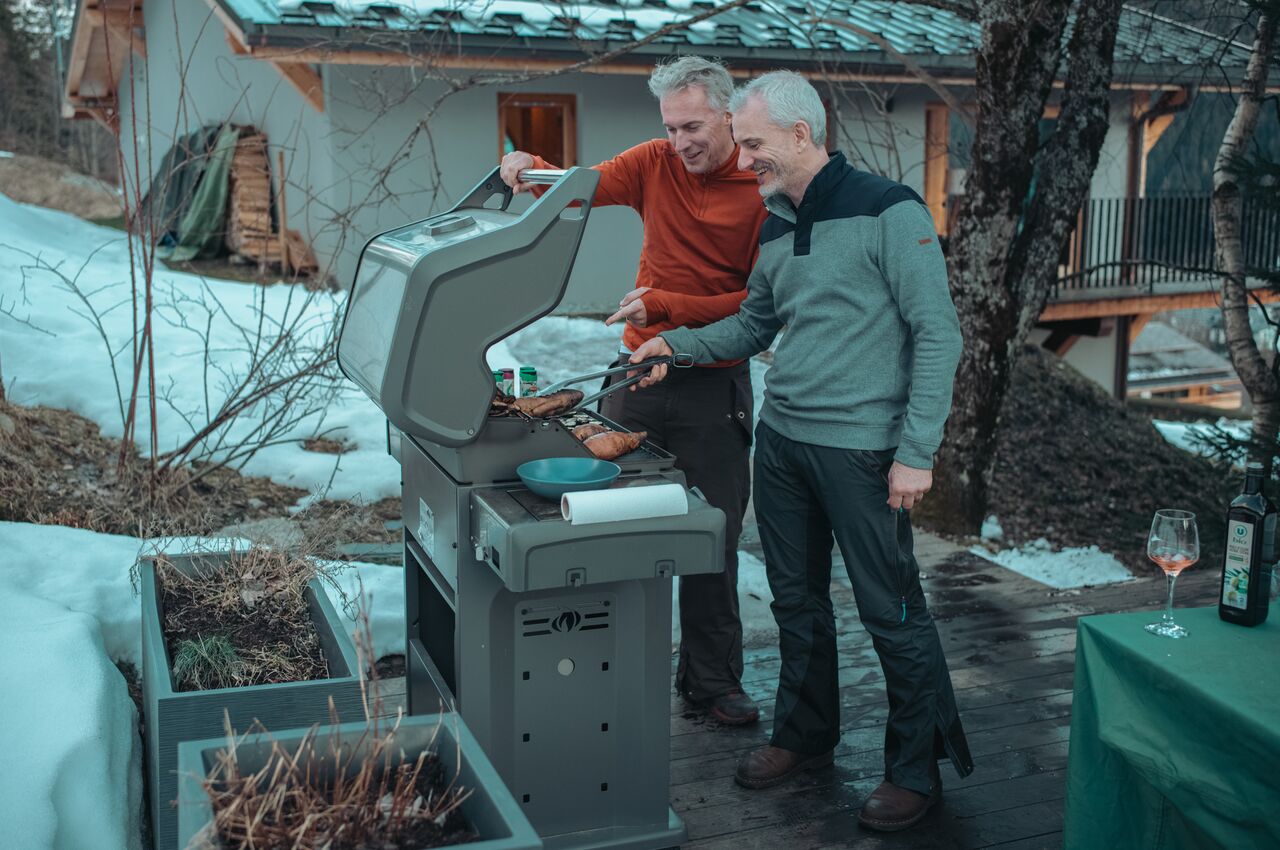 Two men stand by a gas grill, smiling as they turn steaks and sausages with tongs during a winter barbecue on a snowy chalet deck.
