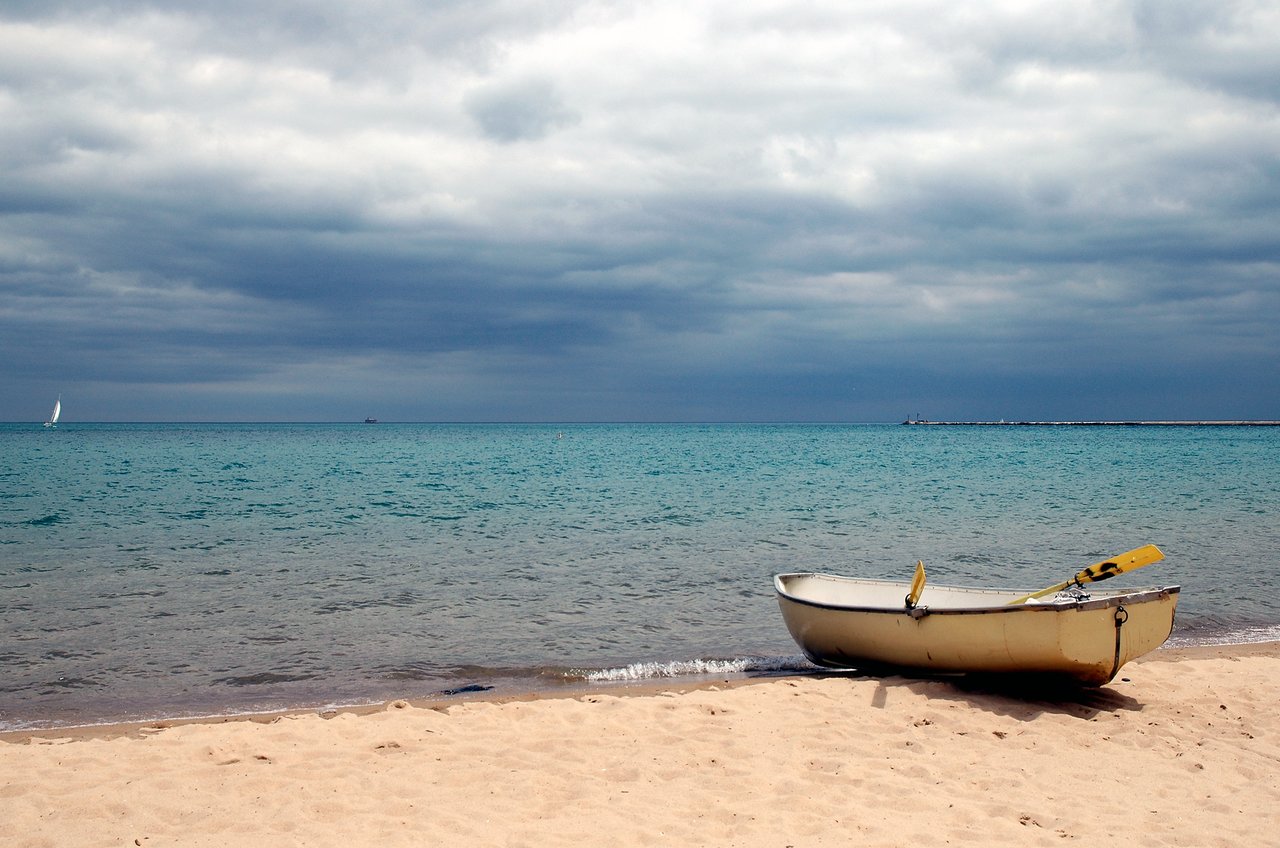 A small yellow boat with oars rests on the sandy shore near calm water under a cloudy sky.