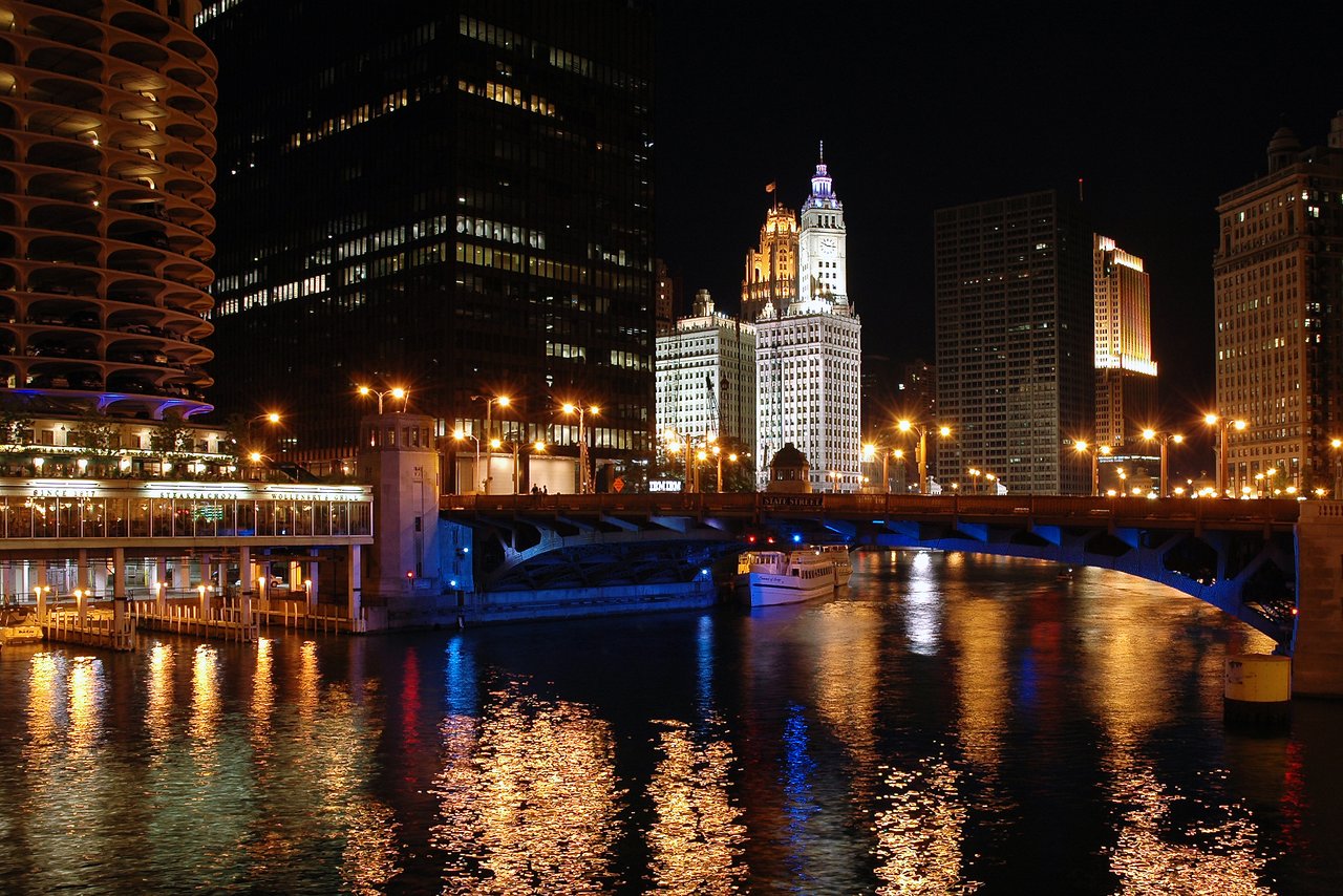 A nighttime view of Chicago with illuminated buildings, a bridge, and reflections on the river.