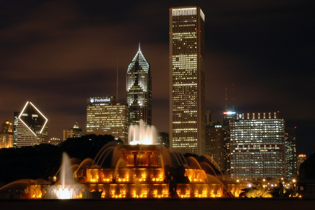 A brightly lit fountain with water jets in front of Chicago's illuminated skyline at night.