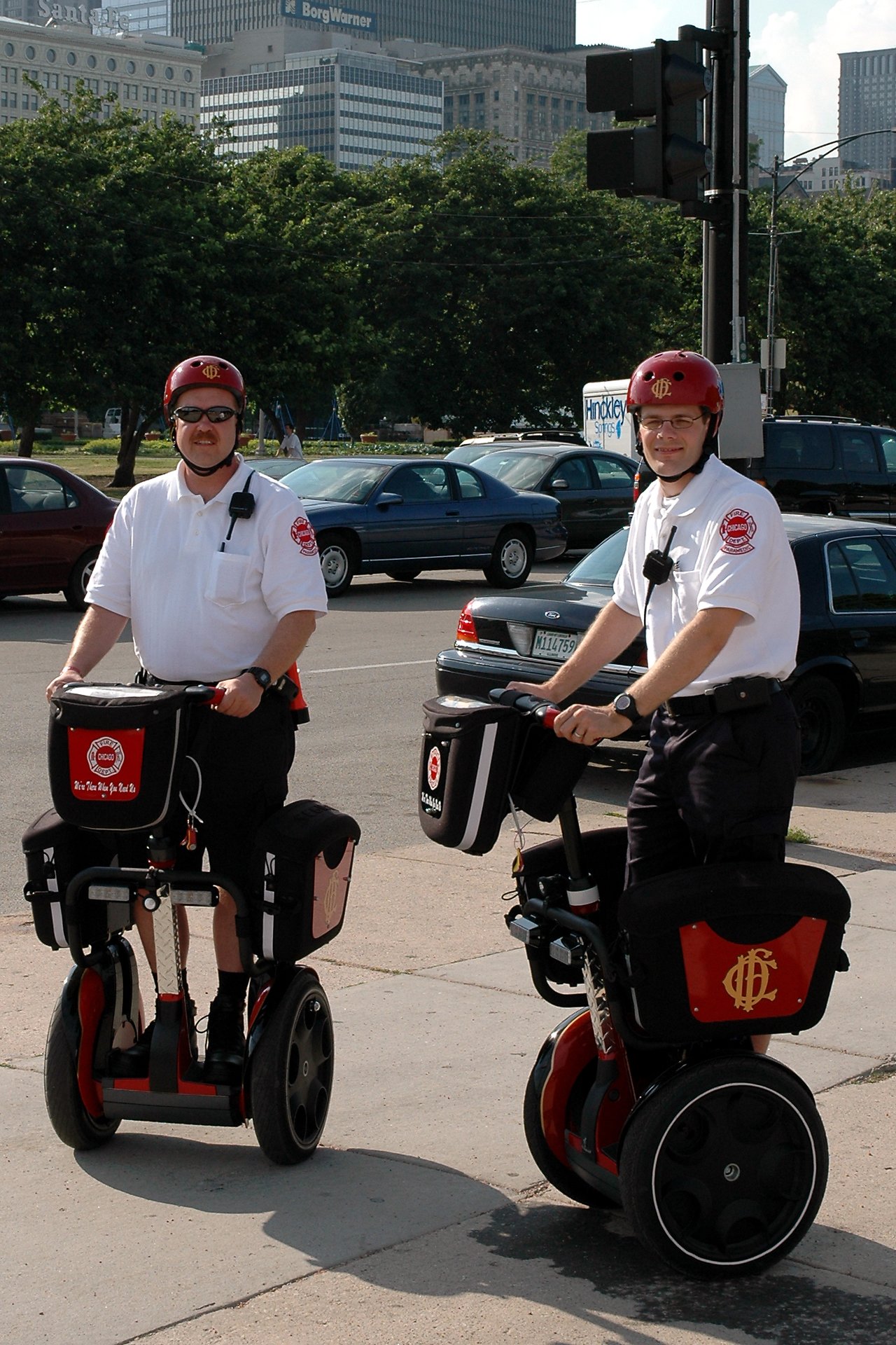 Two firefighters in uniform ride Segways on a sidewalk, wearing helmets and radios, with a cityscape in the background.
