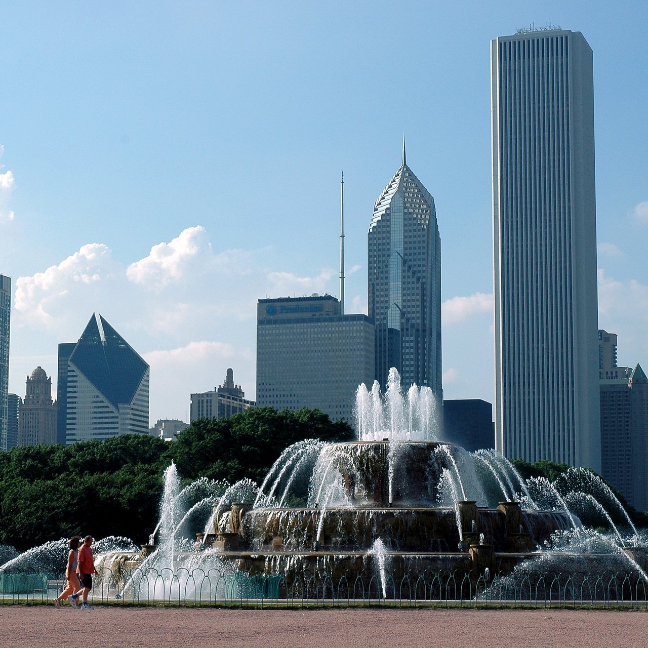 A large fountain with multiple water jets spraying, with two people walking nearby and tall buildings in the background.