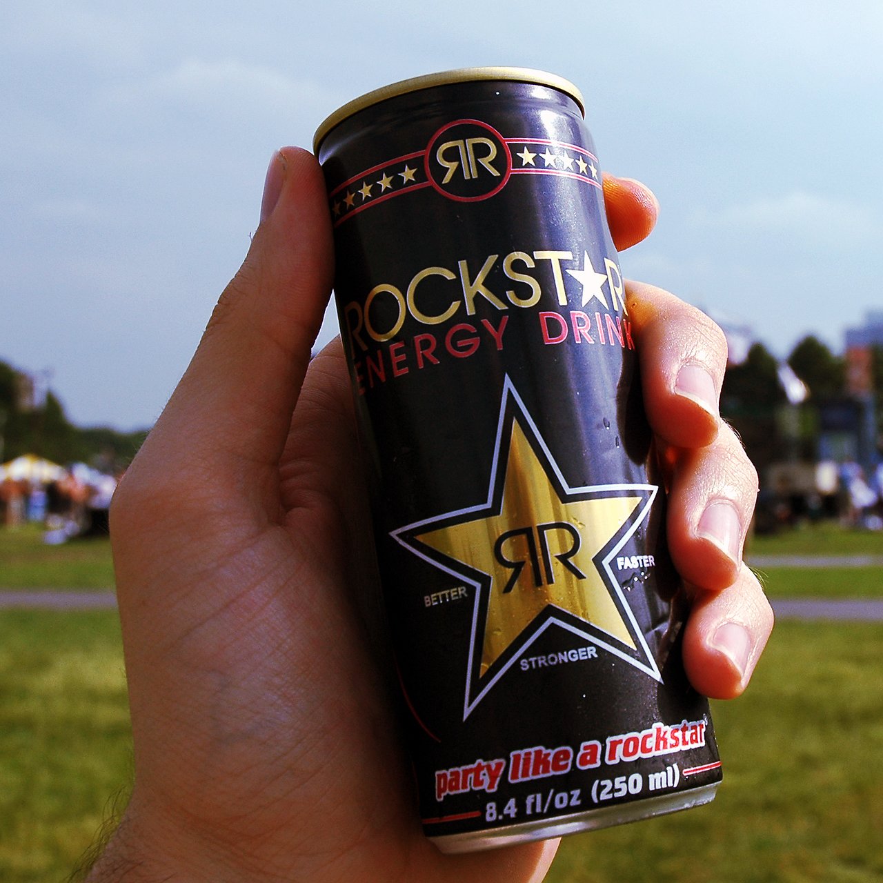 A hand holds a Rockstar Energy Drink can outdoors with a blurred background of grass and people.