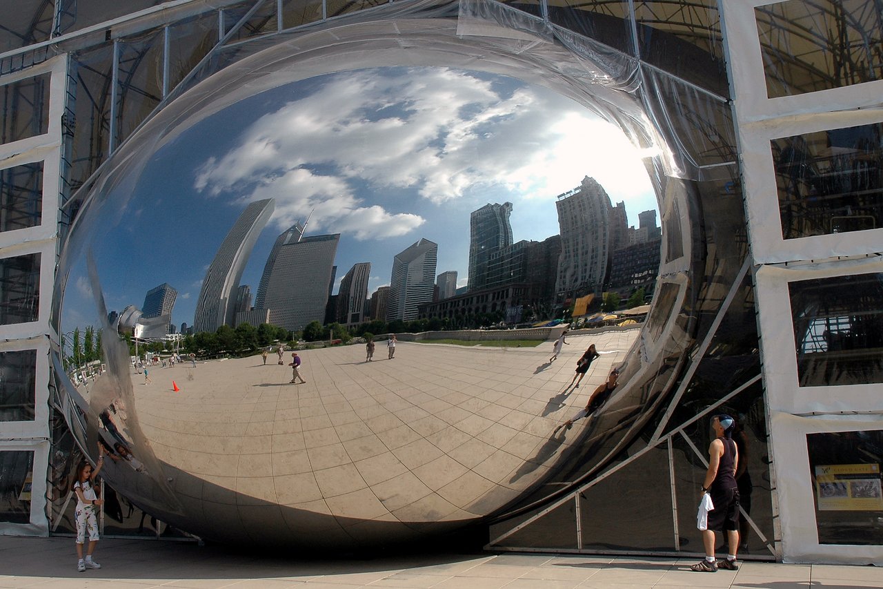 A person takes a self-portrait in the reflective surface of Chicago's Cloud Gate sculpture, with city buildings visible.