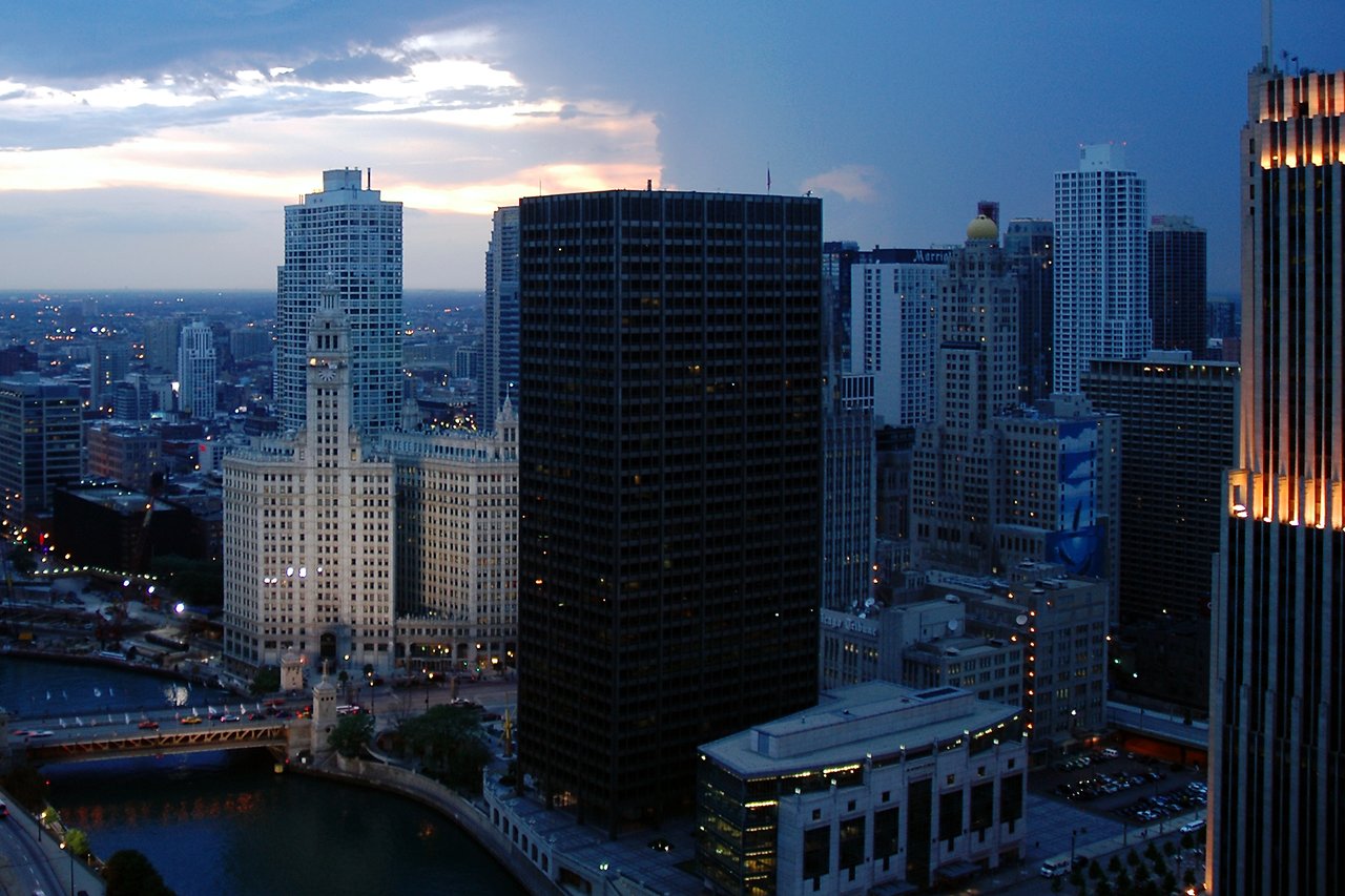 A city skyline view from a high-rise room, showing tall buildings, a river, and a bridge at dusk.