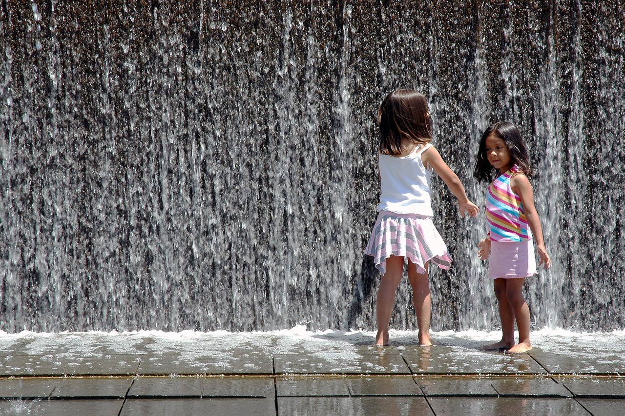 Two young girls play barefoot in front of a cascading water wall, splashing and enjoying the water.
