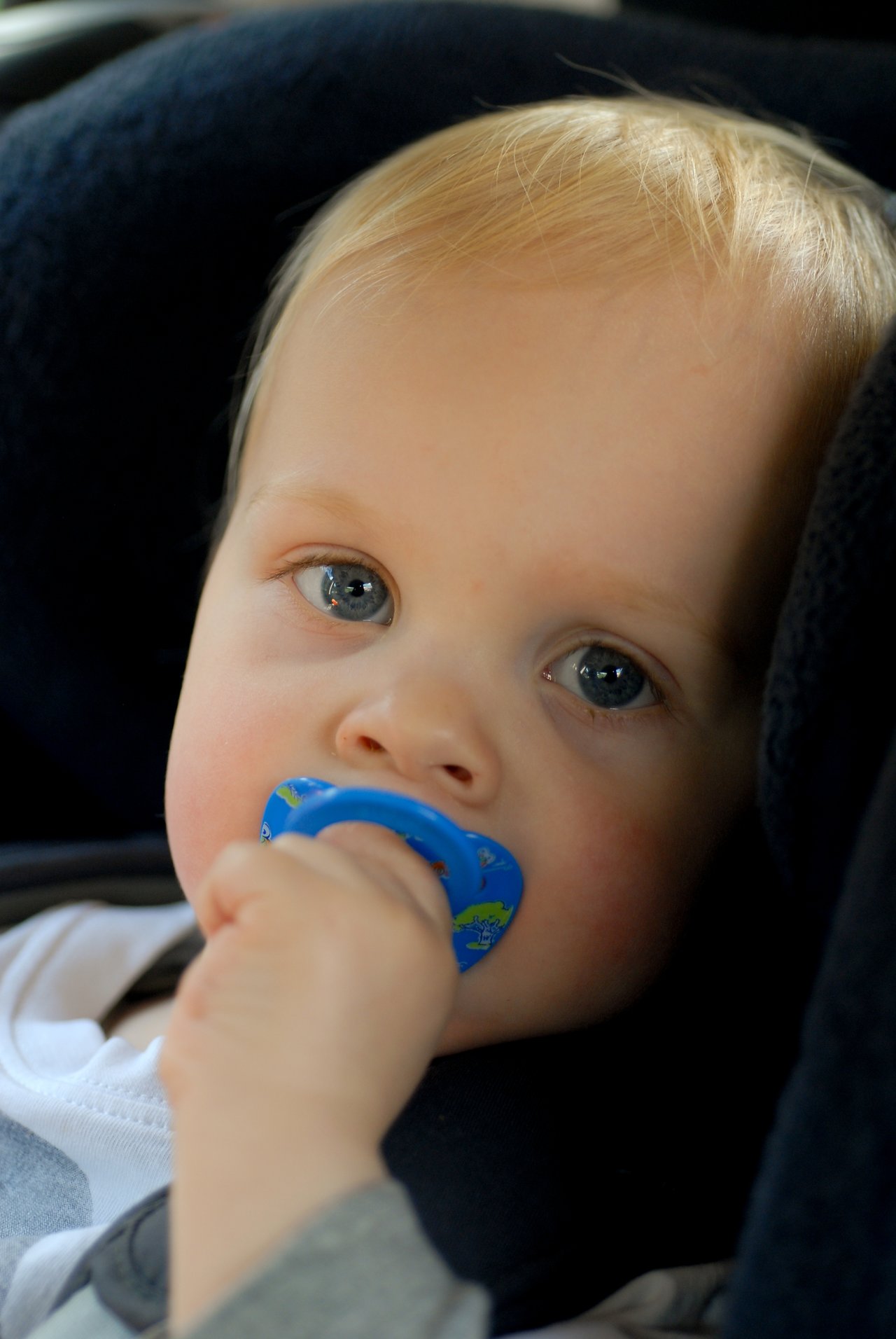 A baby with a blue pacifier sits in a car seat, looking ahead.