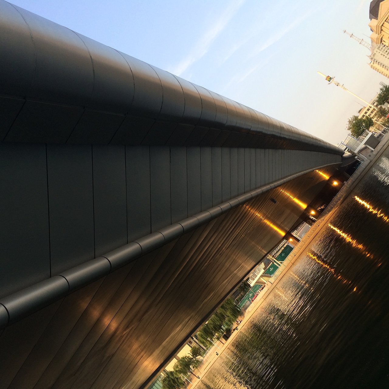 A modern bridge with metallic panels reflecting lights, spanning over a calm body of water at dusk.