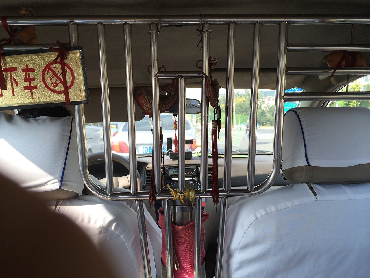 Metal security bars separate the driver and passengers inside a taxi, with a no-exit sign and hanging decorations.