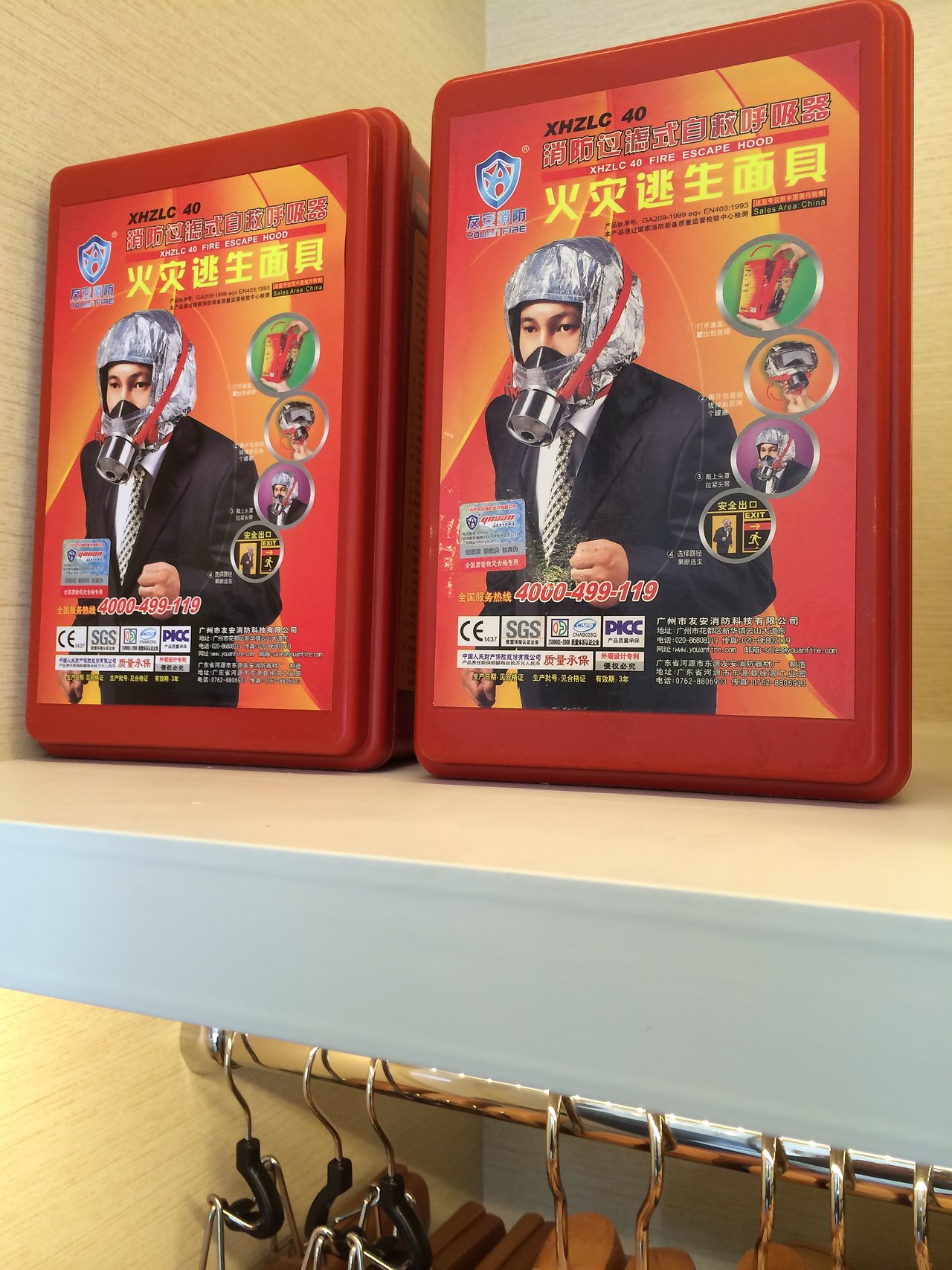 Two red fire escape hood kits are placed on a shelf above a clothing rack with hangers.