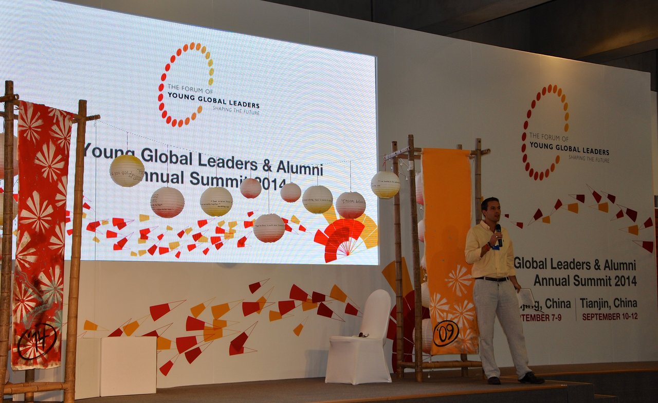 A speaker in a yellow shirt holds a microphone on stage at the Young Global Leaders Summit 2014.