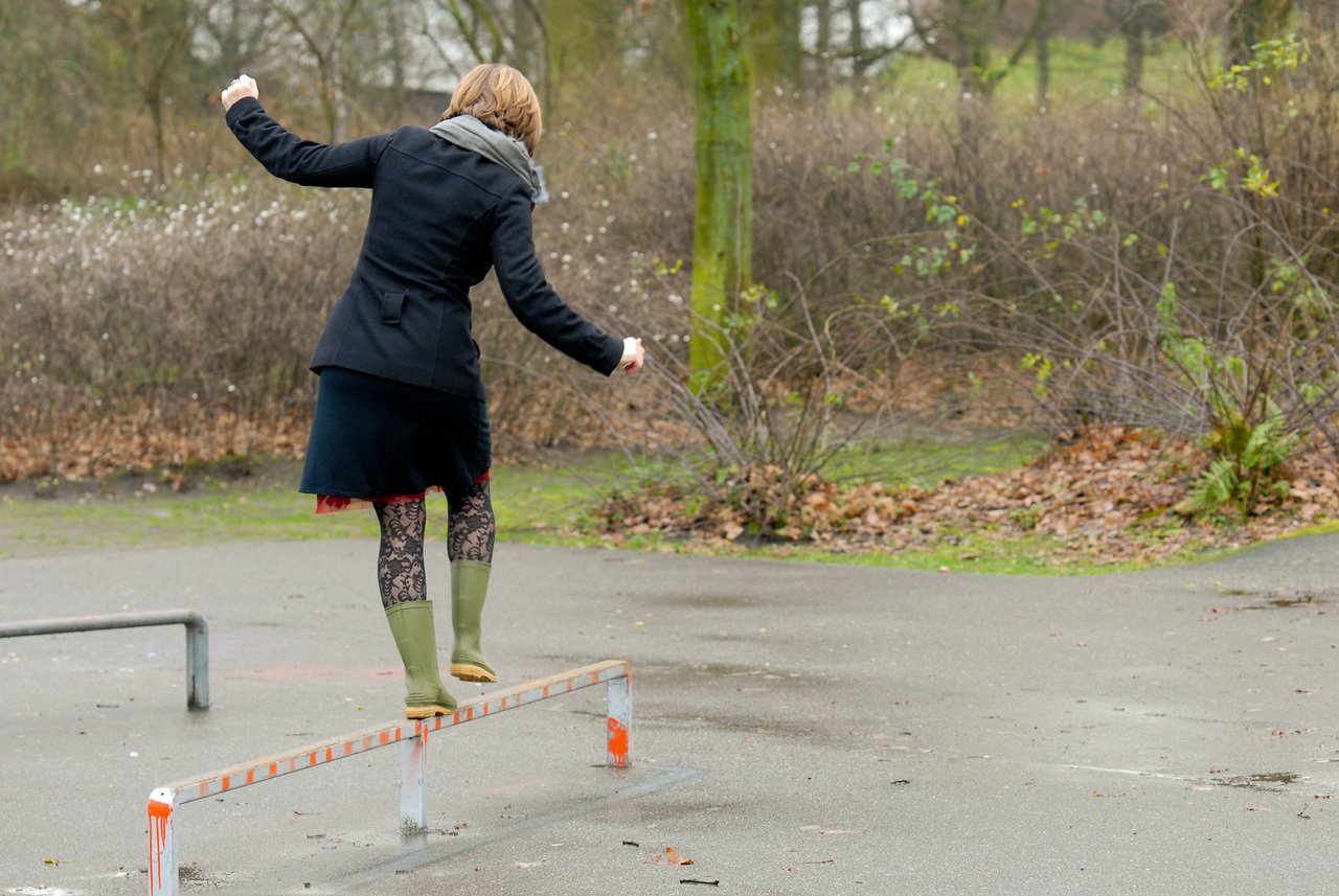 A person in a coat and boots balances on a metal rail in an outdoor park.