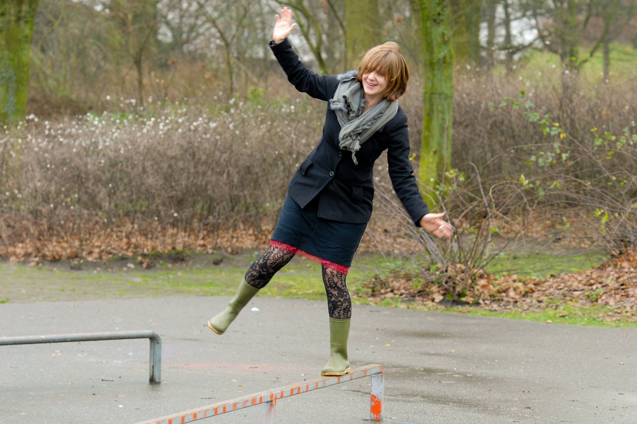 A person in a black coat and green boots balances on a narrow rail outdoors, arms outstretched for stability.