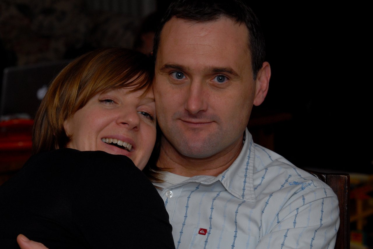 A woman smiles while hugging a man in a white shirt, who looks directly at the camera.